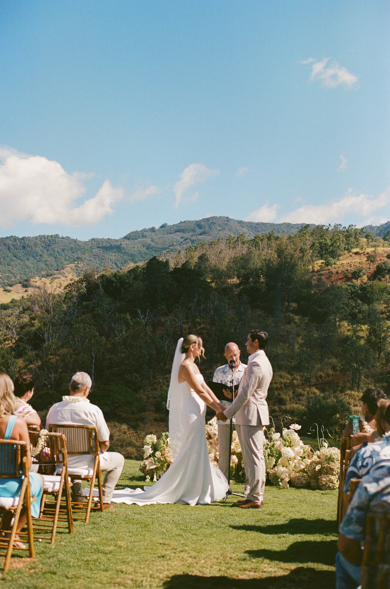 The bride and groom hold hands during their outdoor ceremony with an officiant in front of a mountain backdrop at their Hawaii destination wedding.