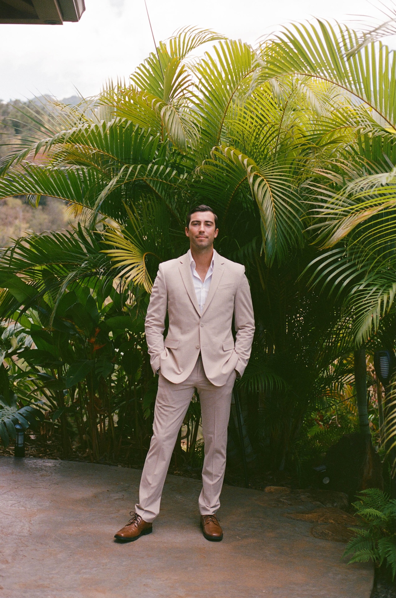 Groom photo with his hands in his pockets standing in front of stunning green palms.
