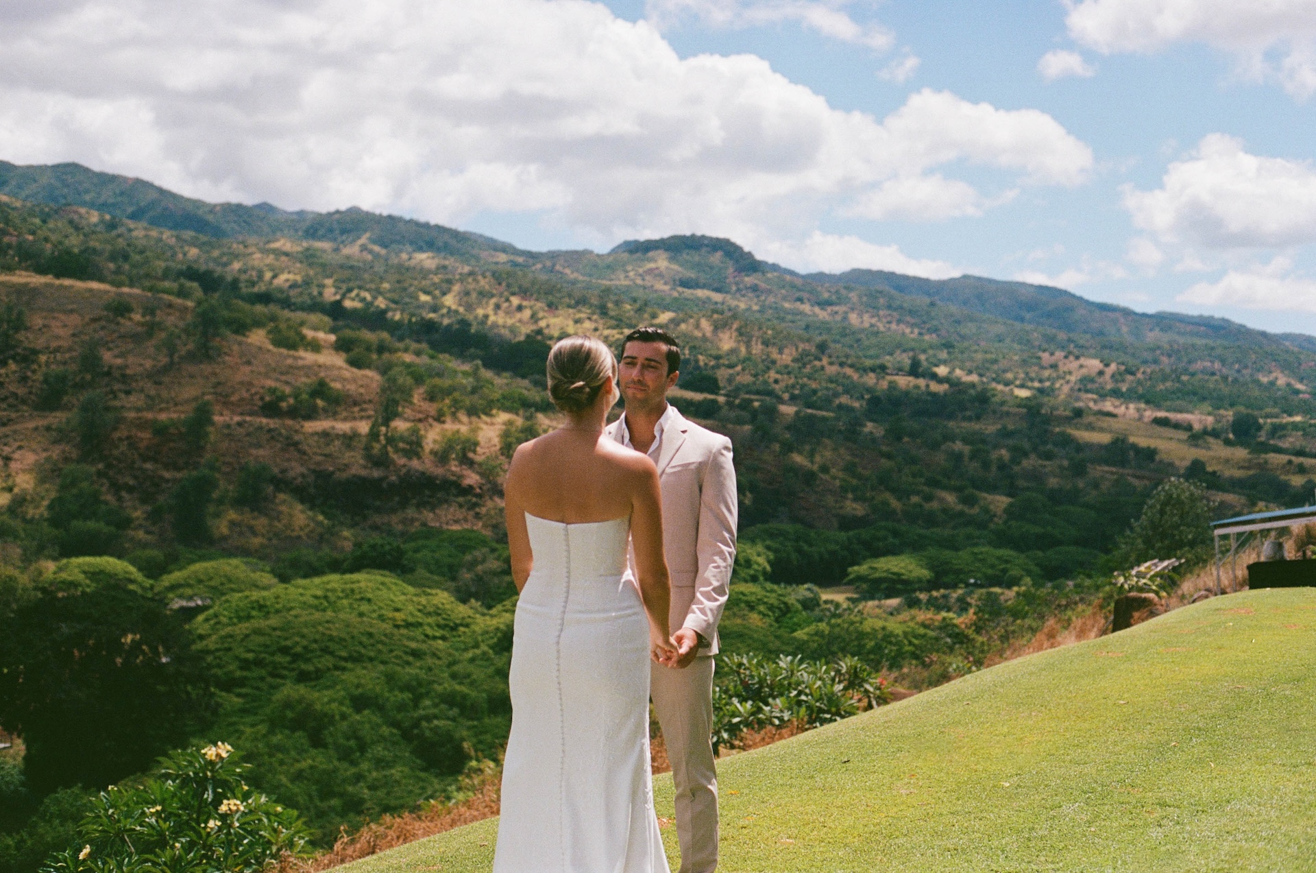 The bride and groom walk hand-in-hand along a grassy hill at golden hour overlooking the Hawaiian landscape.