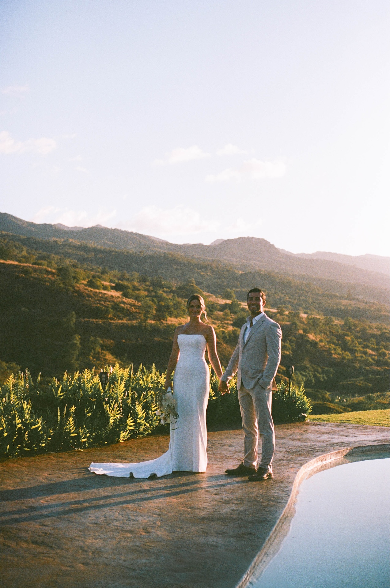 The groom faces the bride as they hold hands on a hillside with mountains in the background under a partly cloudy sky.