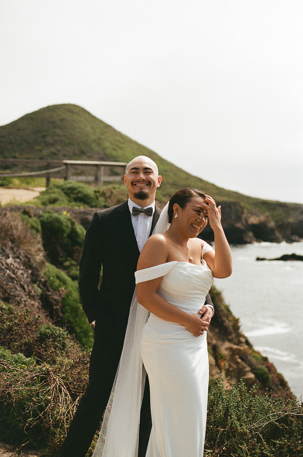 Bride laughing as groom hugs her from behind on a cliffside overlooking the ocean, both smiling during portraits.