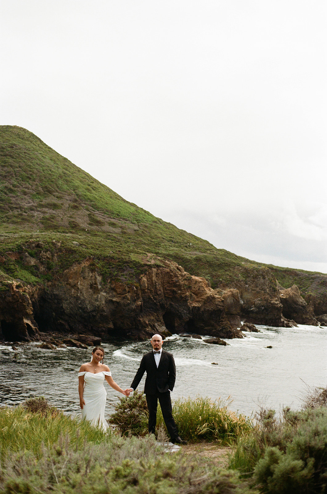 Bride and groom holding hands on a grassy cliff overlooking the ocean and rugged rocky Big Sur coastline.