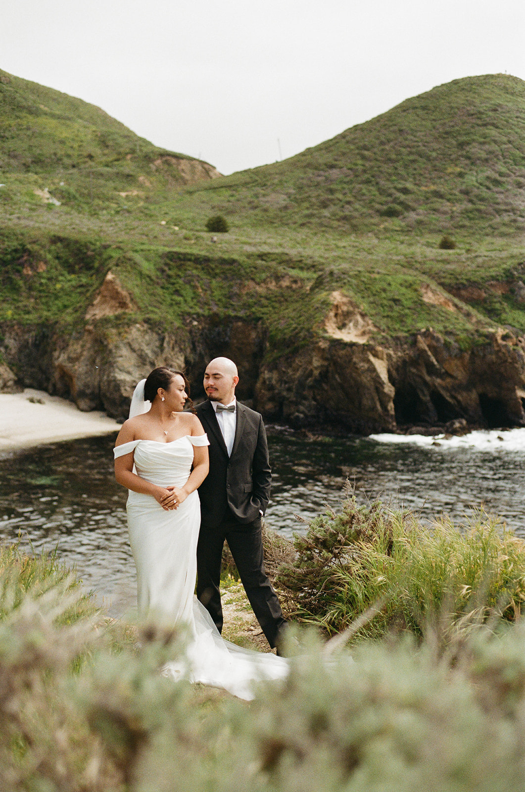 Bride and groom looking at each other on a cliffside, dramatic rocky coastline and green hills in the background.