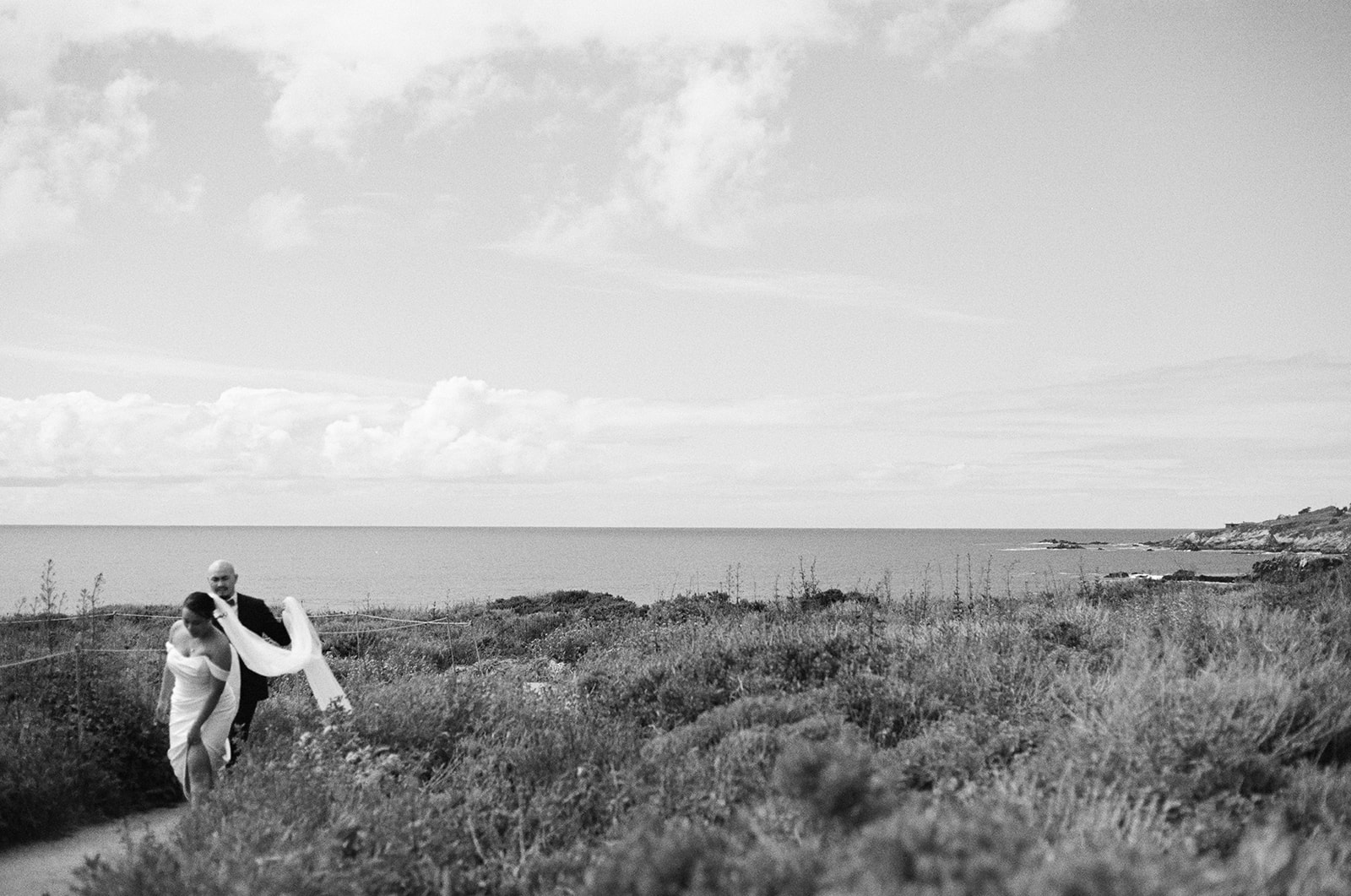 Bride and groom walking along a coastal path with her veil blowing in the wind, ocean and cloudy sky in the background.
