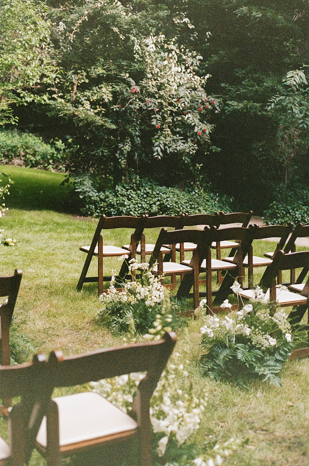 Wooden chairs arranged outdoors in a forest setting with lush greenery and white floral aisle arrangements.