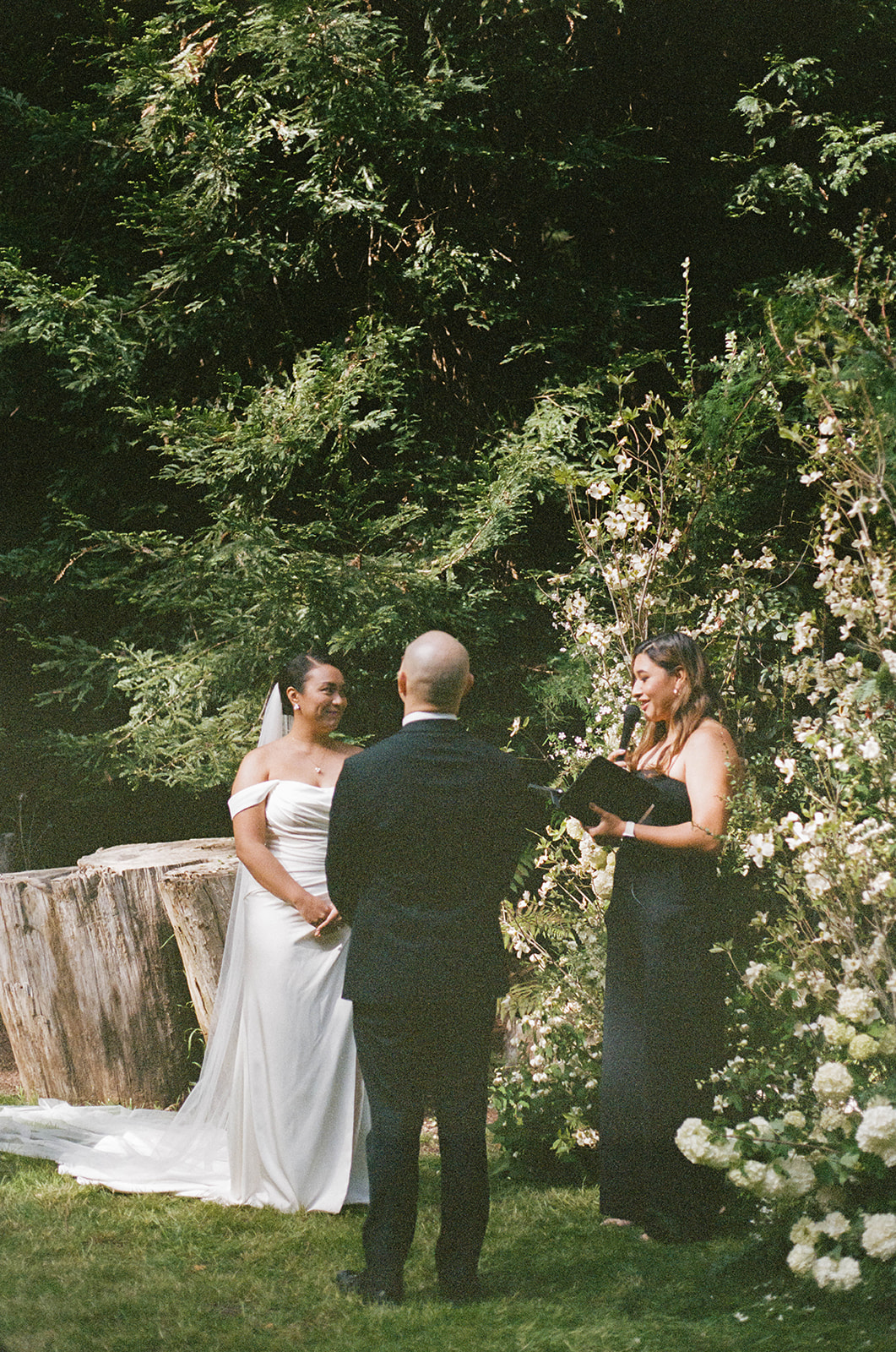 Bride and groom standing with officiant during their forest ceremony, surrounded by blooming white flowers and greenery.