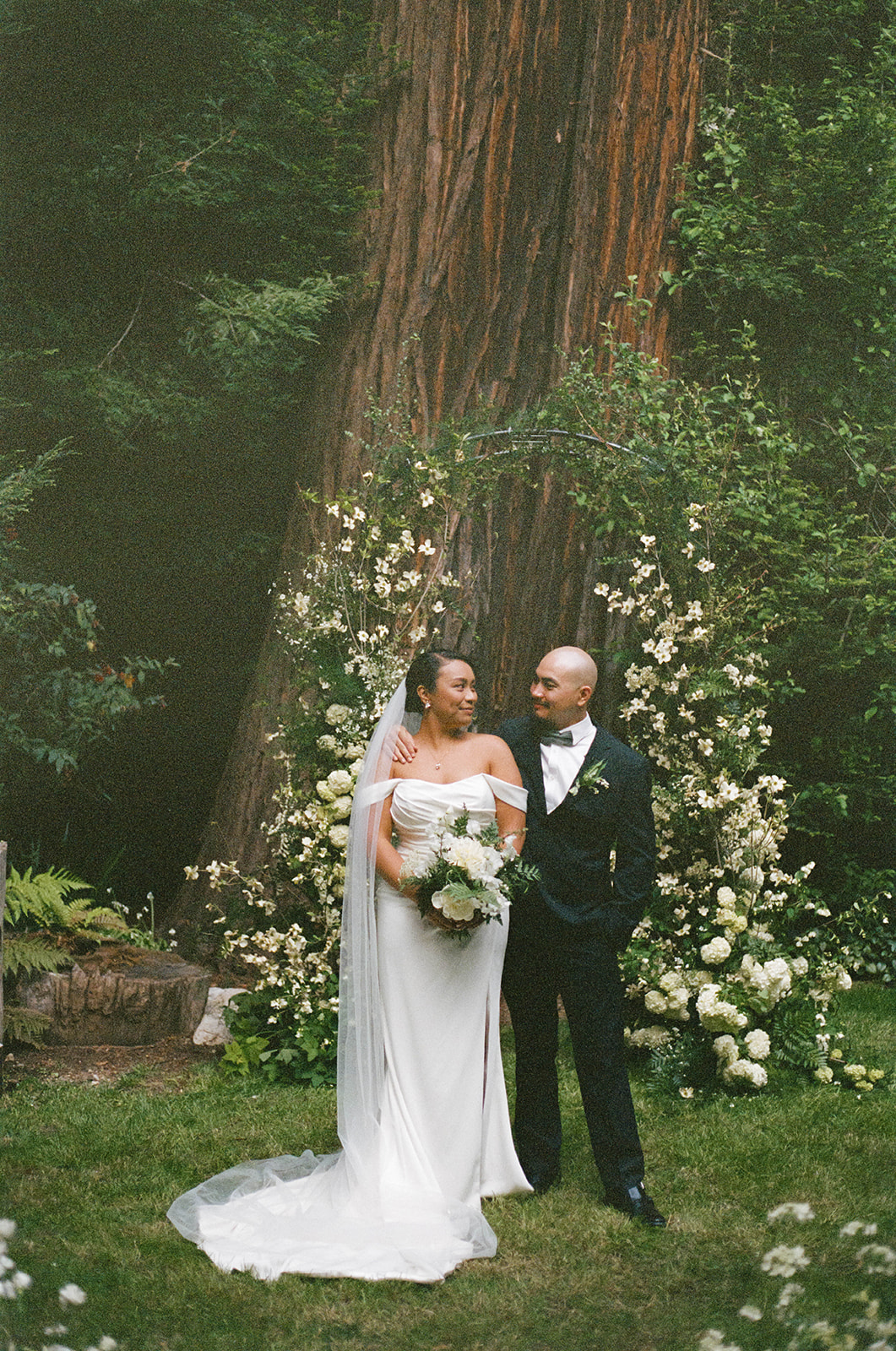 Bride and groom stand together in front of a floral arch under a towering redwood, sharing an intimate moment.