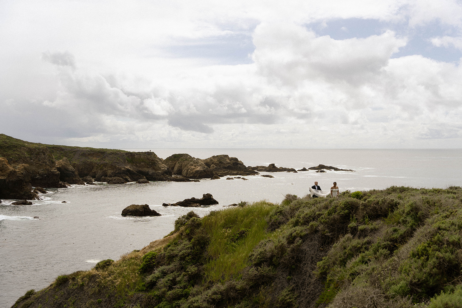 Wide shot of a couple walking near the cliffs by the ocean, carrying a surfboard during their adventurous Big Sur wedding celebration.