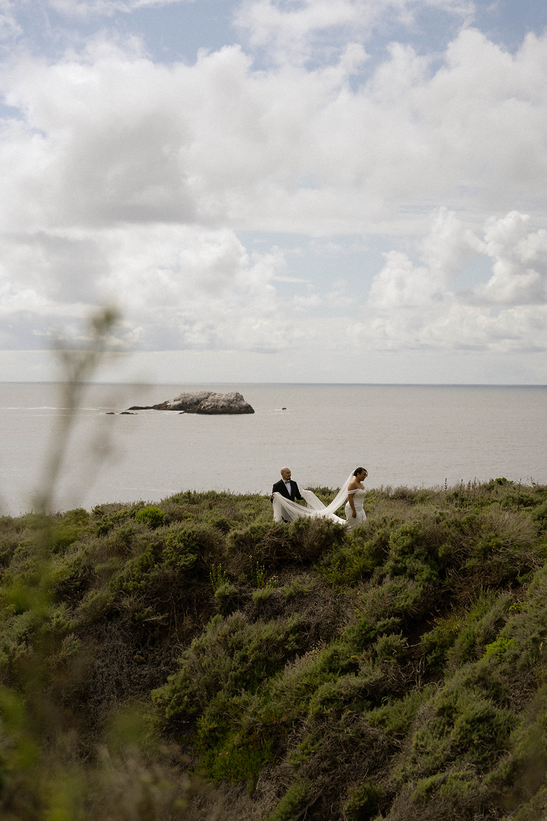 Bride and groom walking along coastal cliffs, with greenery and ocean views surrounding them at their Big Sur wedding.