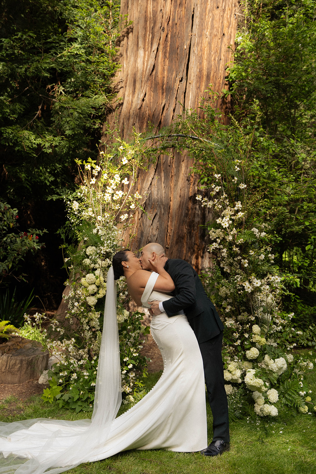 Bride and groom kissing at the altar under a floral arch surrounded by towering trees during their Big Sur wedding.