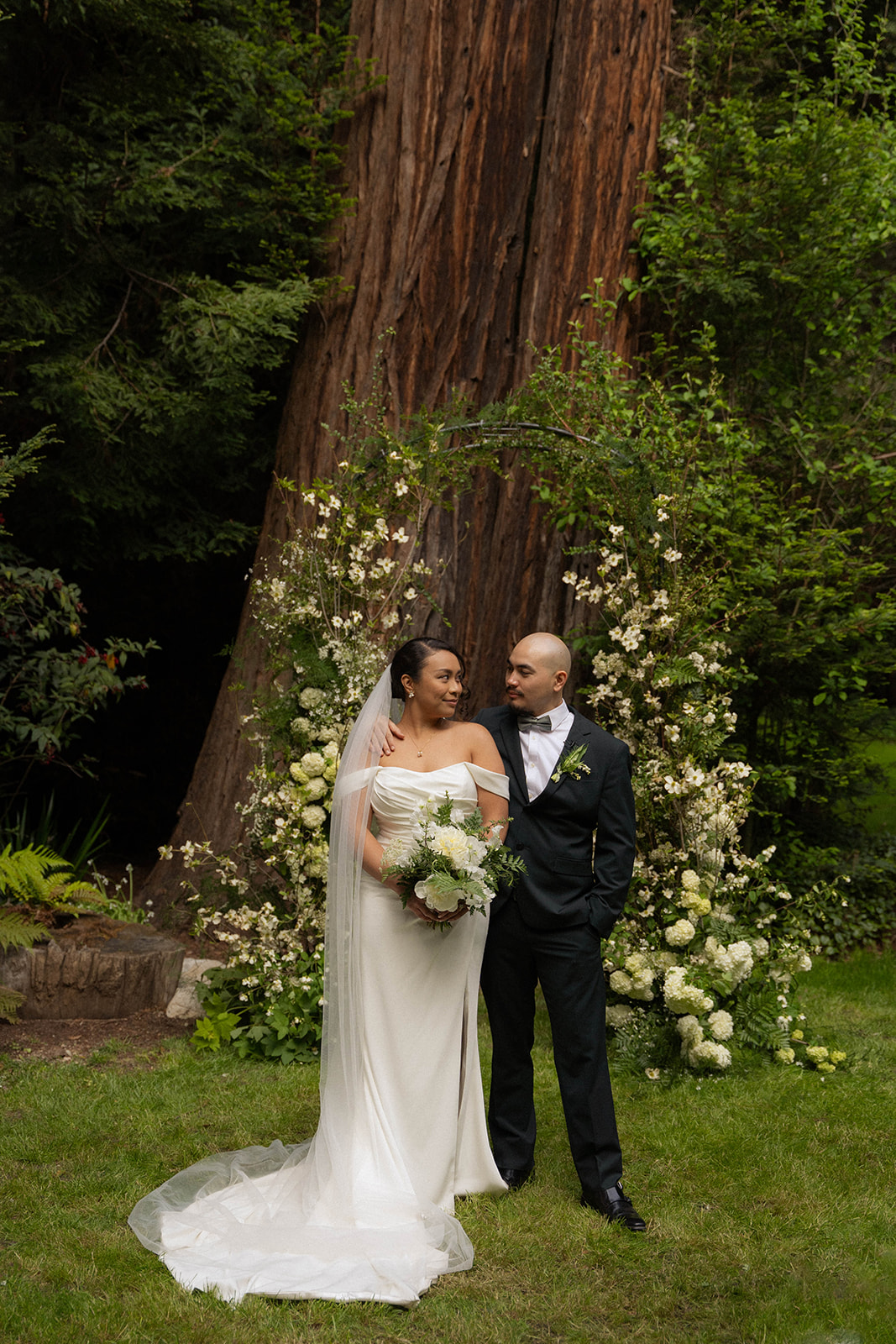 Bride and groom smiling under a floral arch surrounded by redwoods at their intimate Big Sur wedding ceremony.