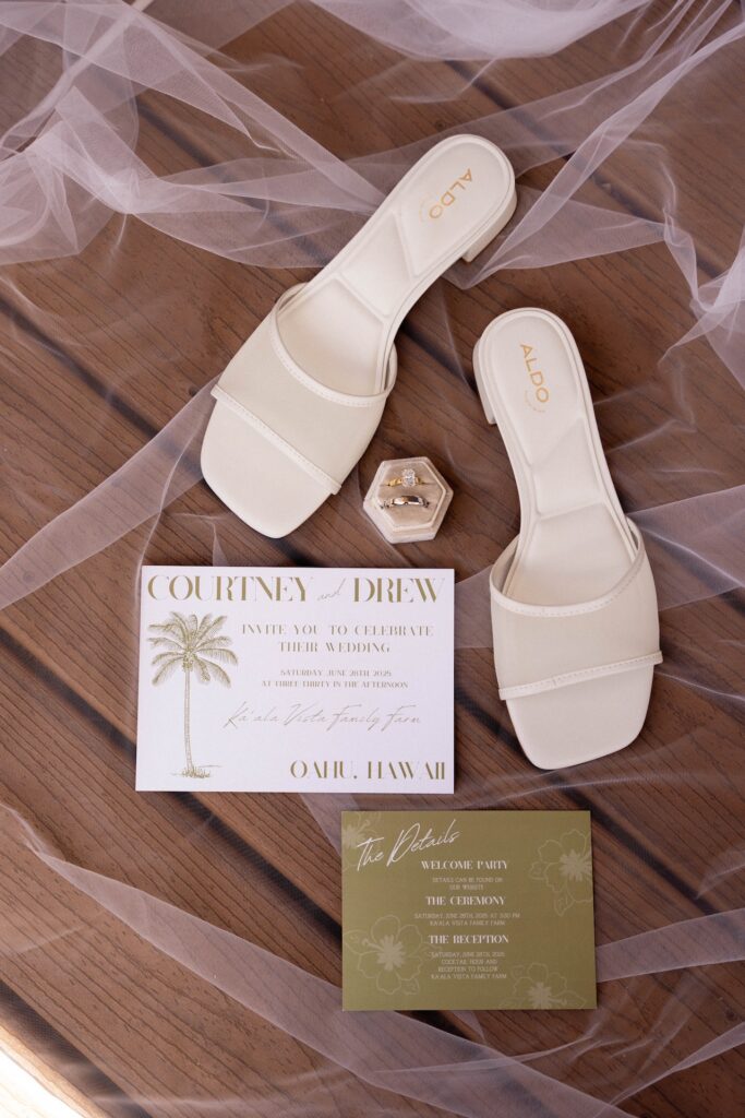 Flat lay of wedding invitations, white bridal shoes, and rings with Oʻahu, Hawaii details included.