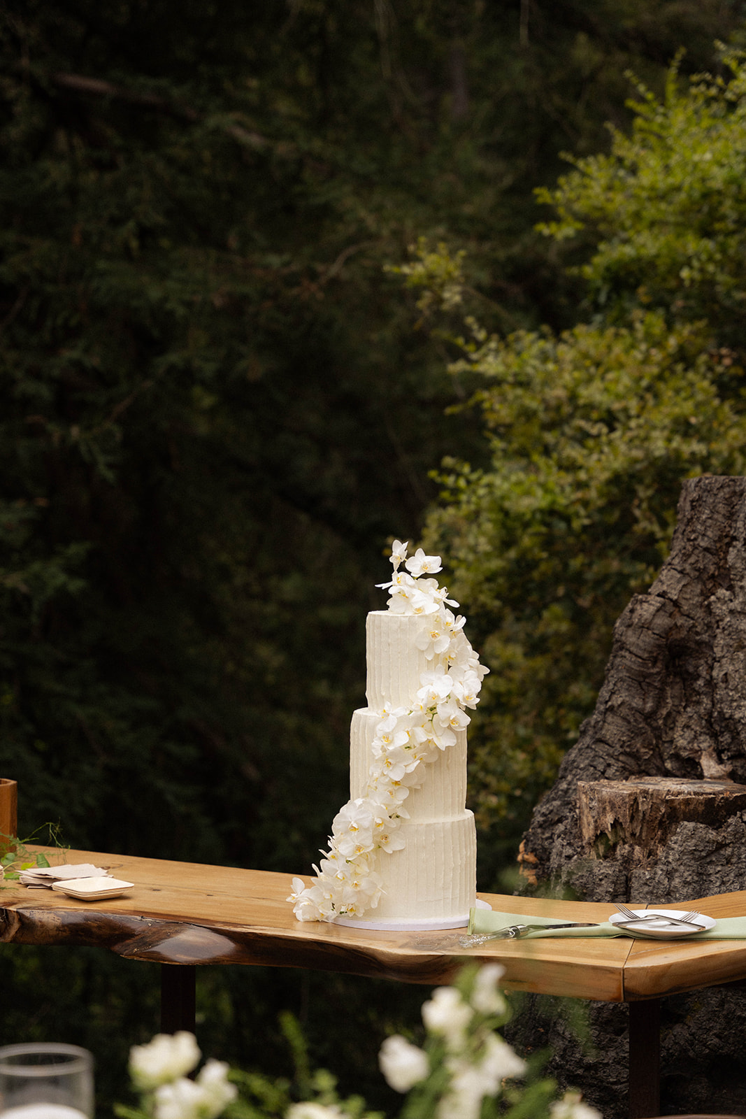 Three-tiered white wedding cake with cascading white flowers set against a forest backdrop at a Big Sur wedding.