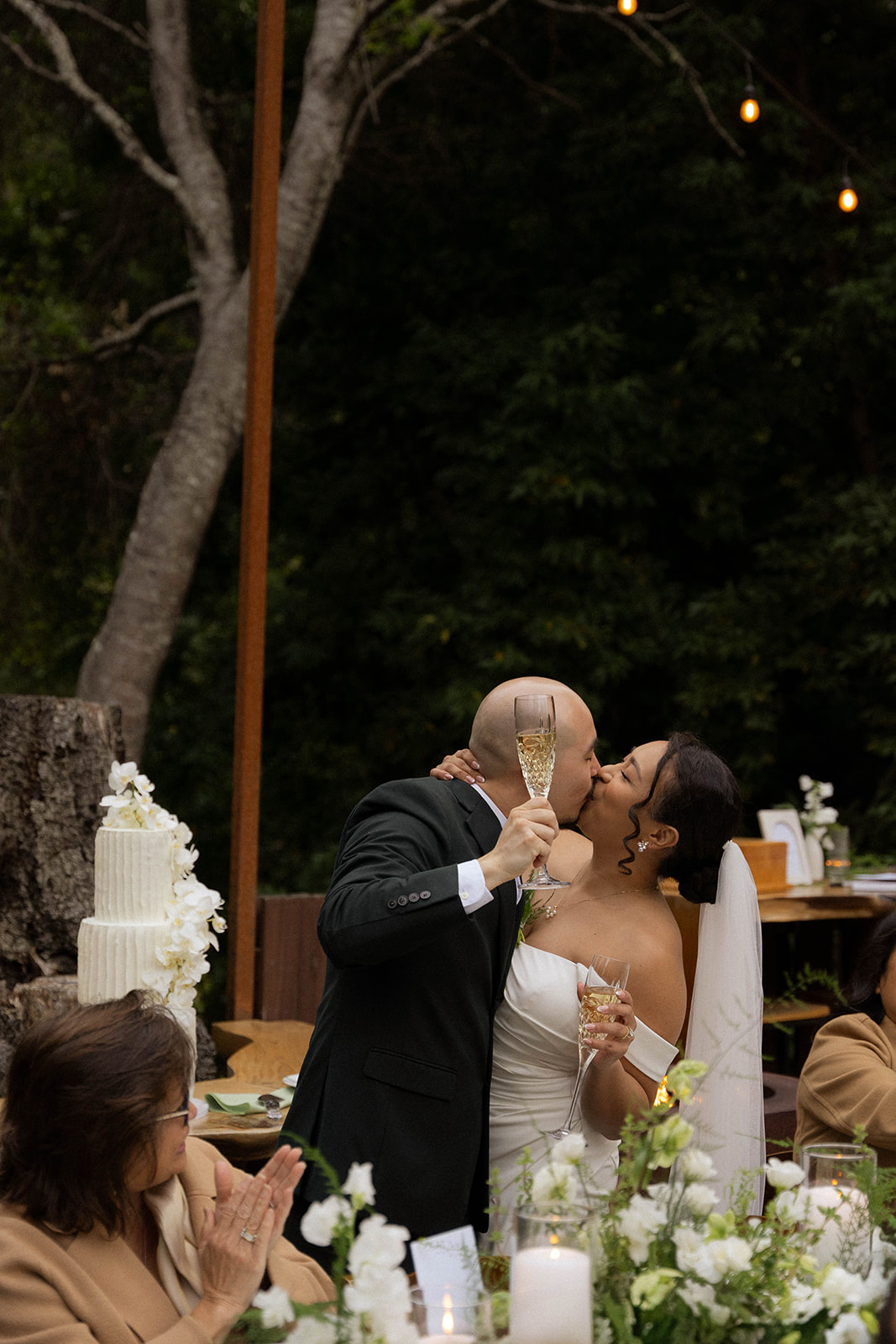 Bride and groom kiss while holding champagne glasses during their Big Sur wedding reception.