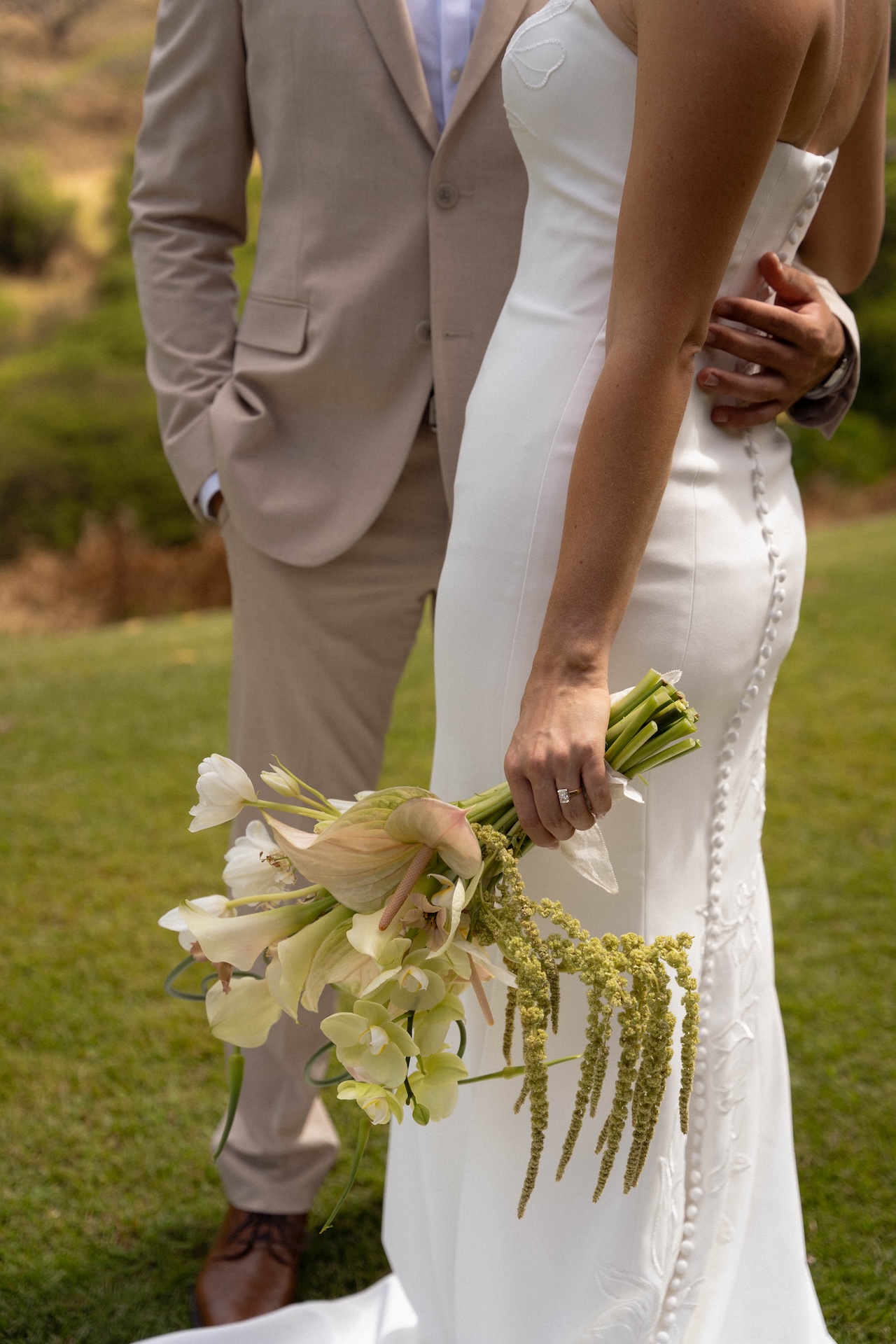 Close-up of the bride’s hand holding a white and green bouquet with the groom’s hand around her waist at Kaʻala Vista.