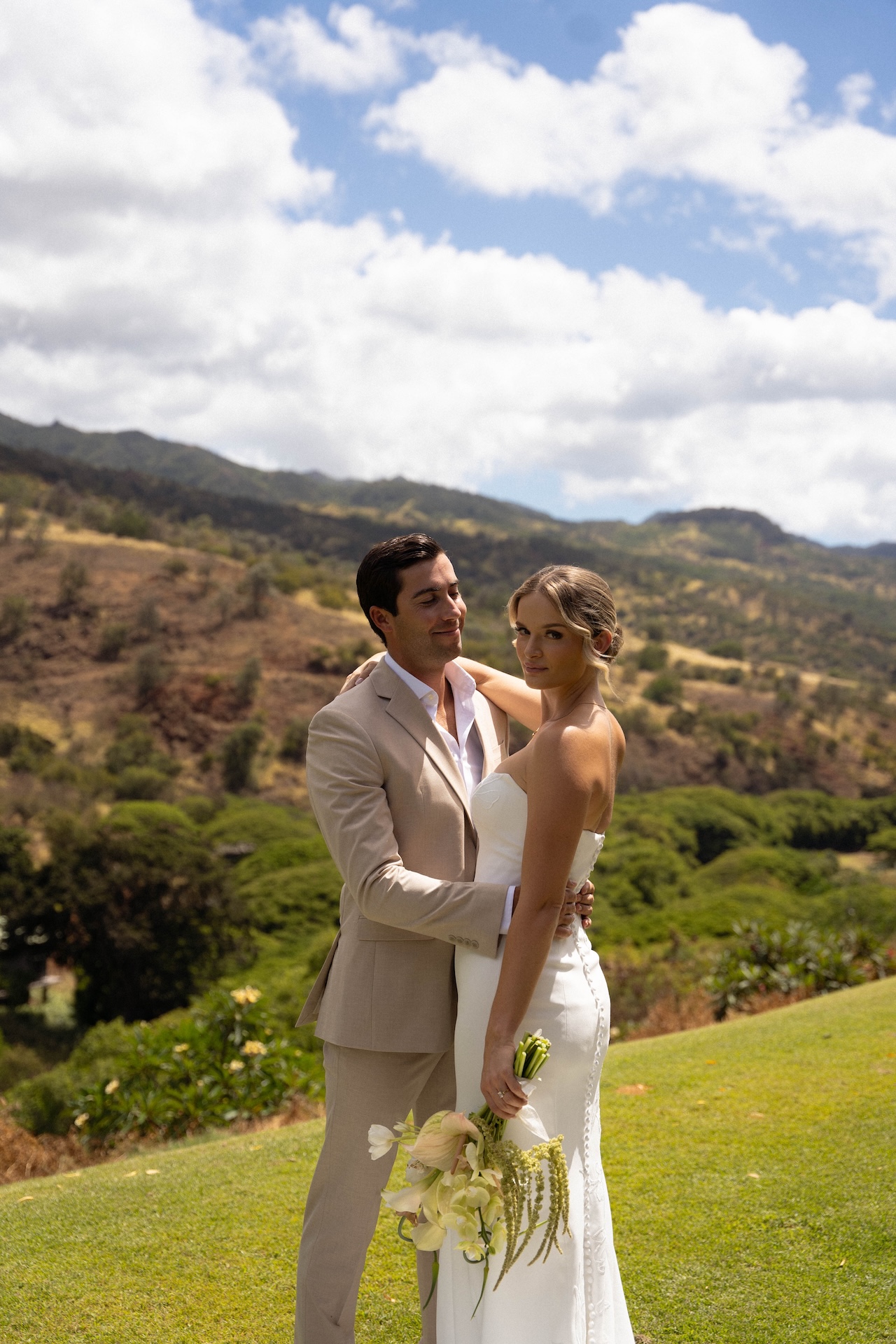 Bride and groom standing on a grassy hill at Kaʻala Vista with North Shore mountains in the background during their Hawaii destination wedding.