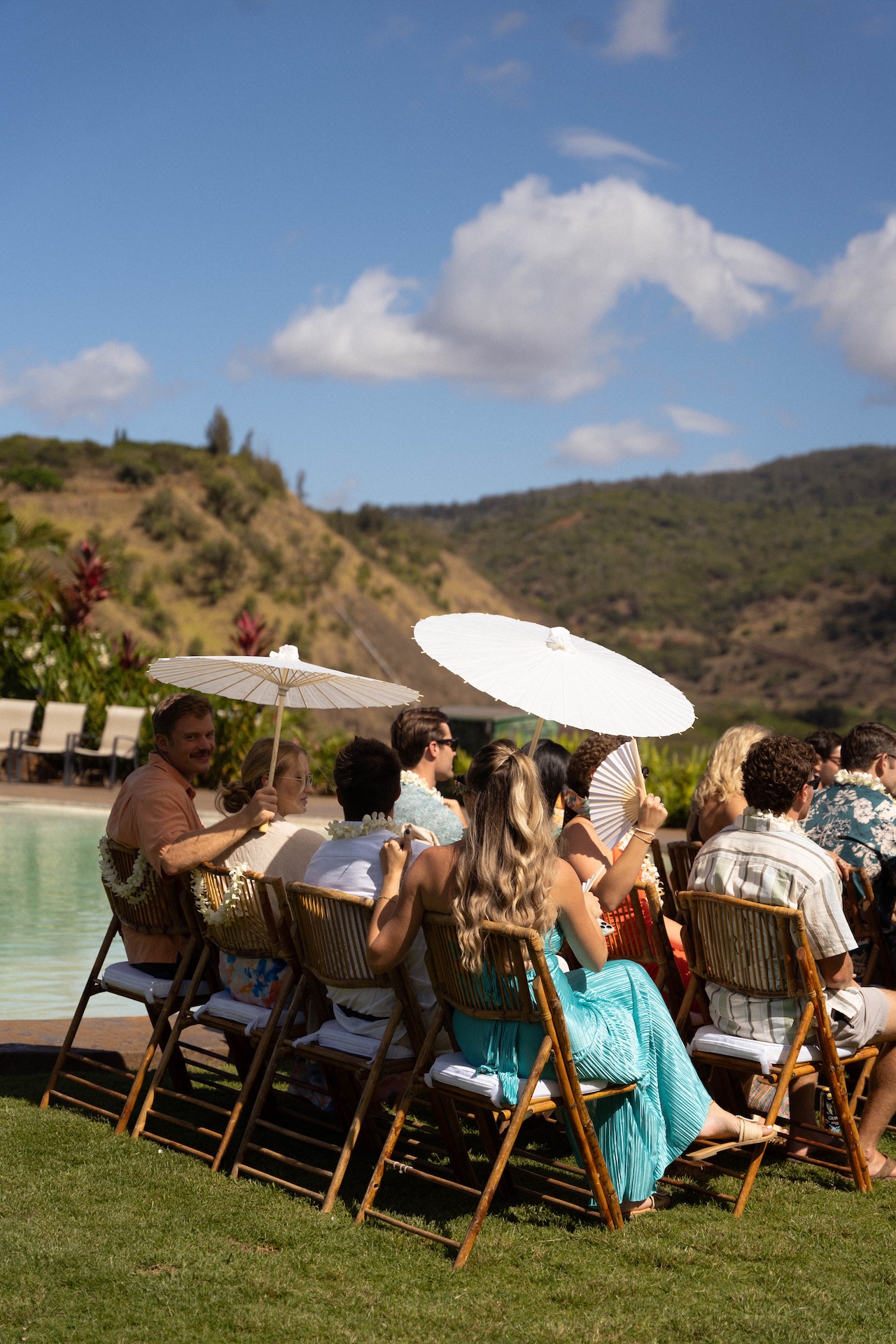 Guests sit in bamboo chairs holding white parasols near the pool before the ceremony begins.

