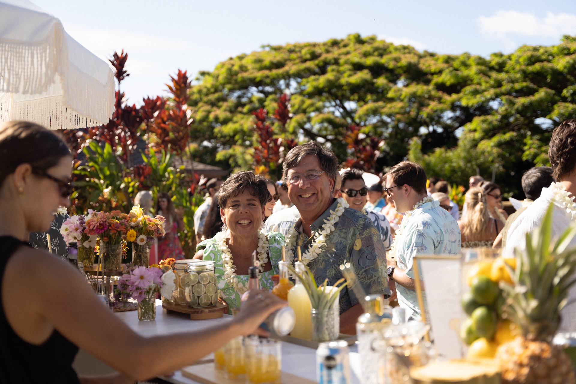 Guests wearing leis stand at an outdoor bar decorated with flowers while drinks are being prepared.