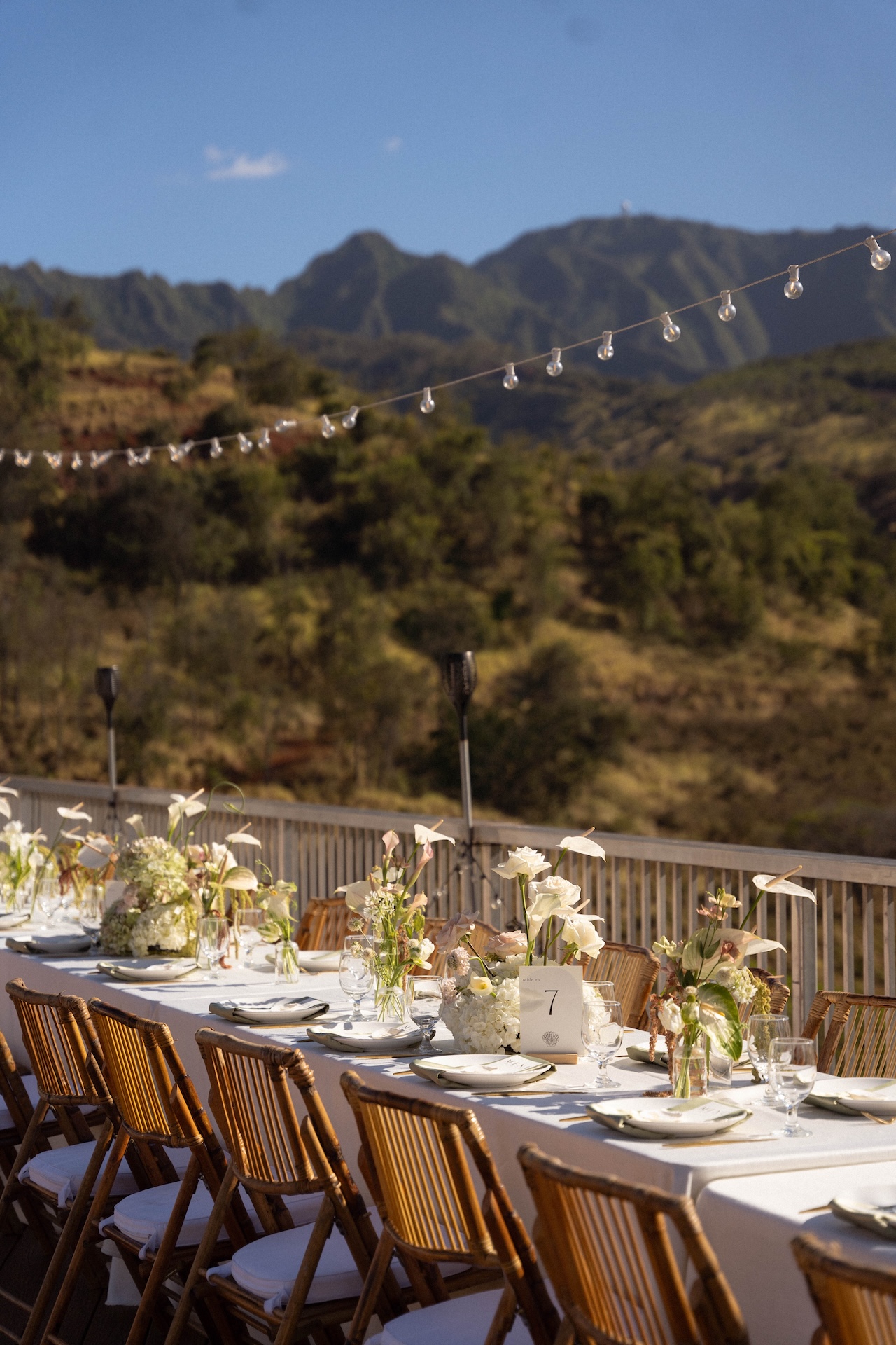 A long table setup for the reception with white and green flowers, table numbers, candles, and fairy lights overlooking the mountains.