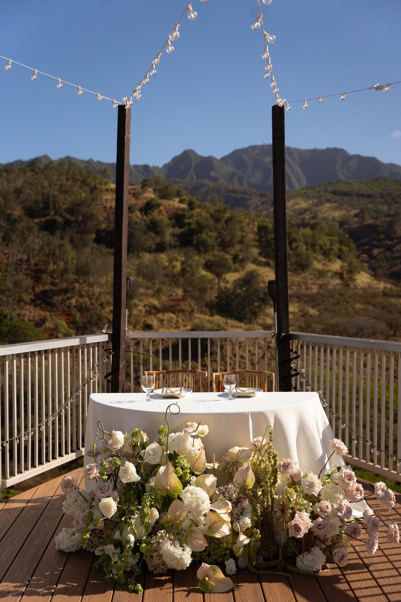 A sweetheart table with white linens and abundant floral arrangements sits on a deck overlooking mountains.