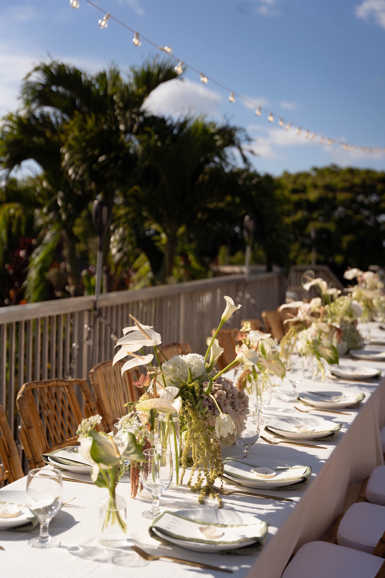 A long reception table with white linens, floral arrangements, and rattan chairs.

