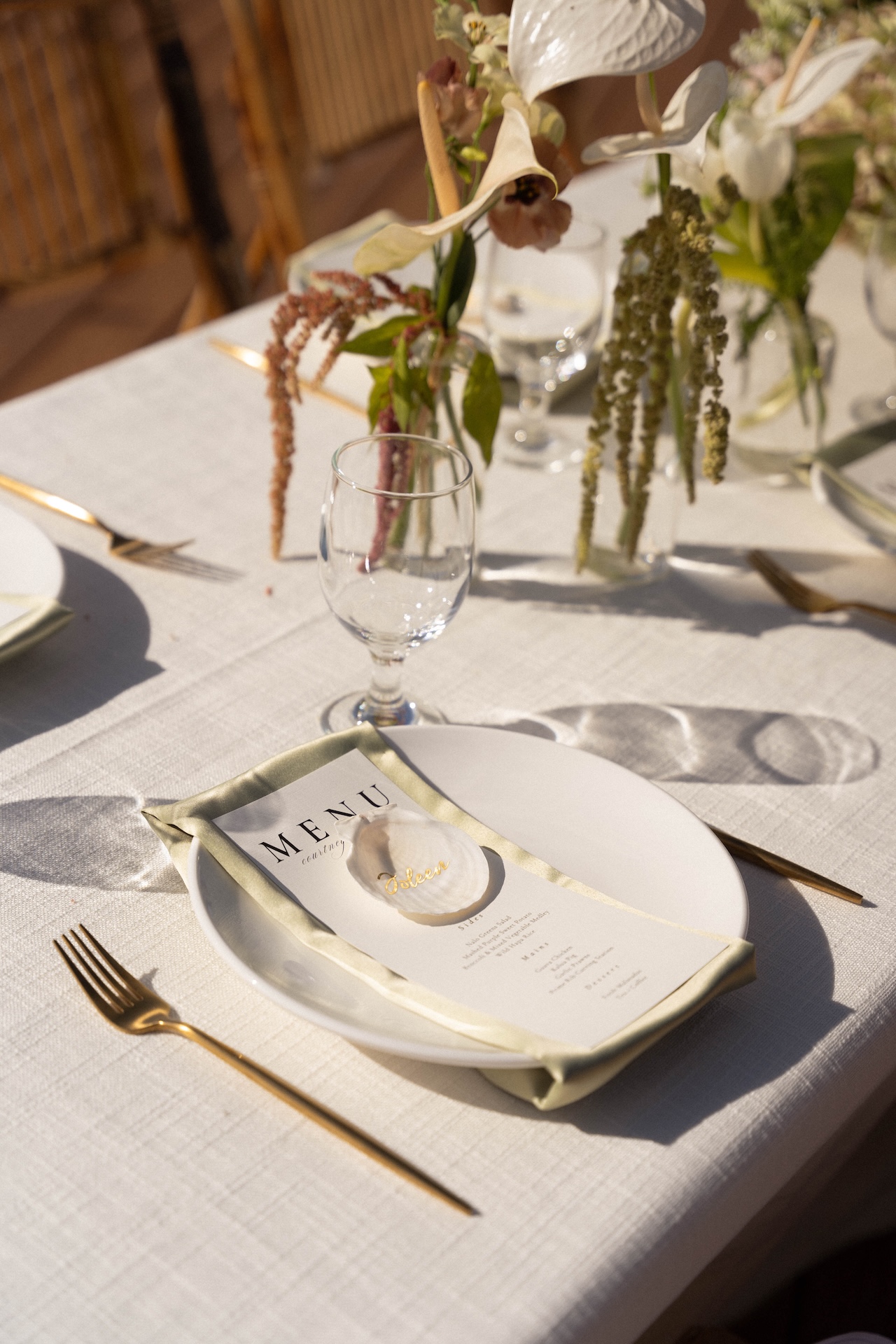 A white plate with a printed menu, gold flatware, and a shell name card on a linen tablecloth.

