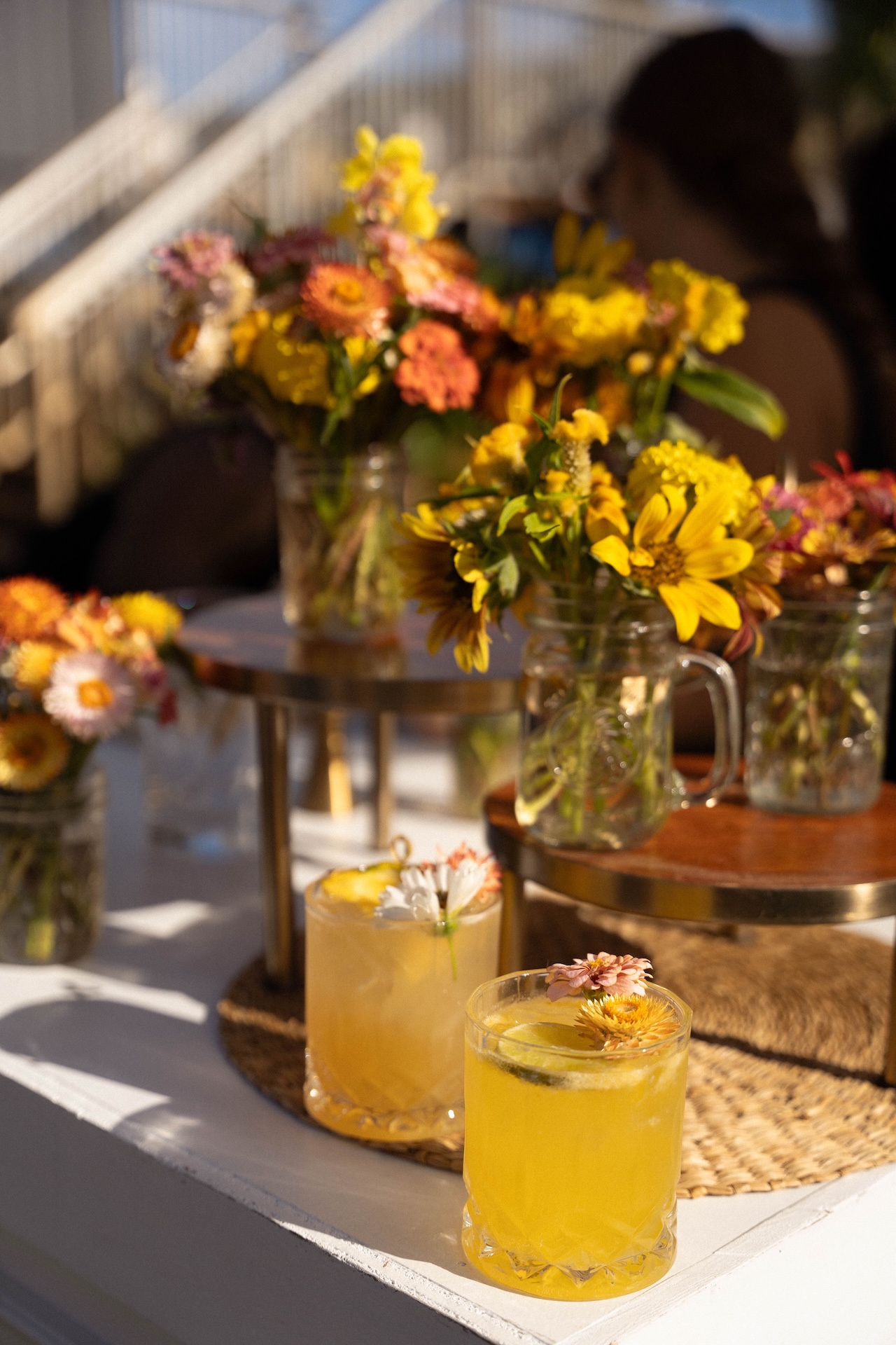 Two yellow cocktails with flower garnishes sit on a bar surrounded by jars of bright flowers.