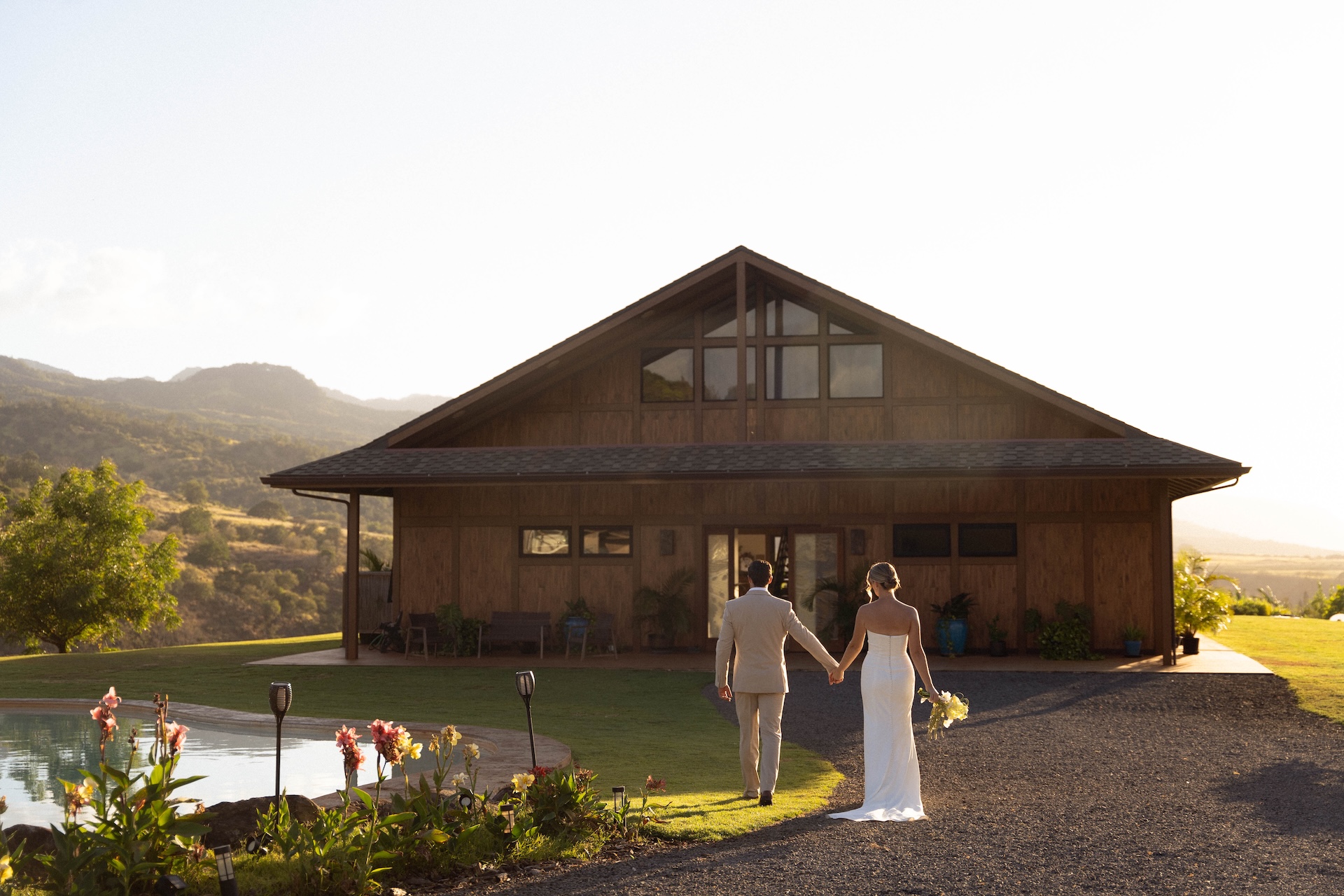 Bride and groom walking hand in hand toward the Kaʻala Vista venue with the pool and mountains in view at sunset.