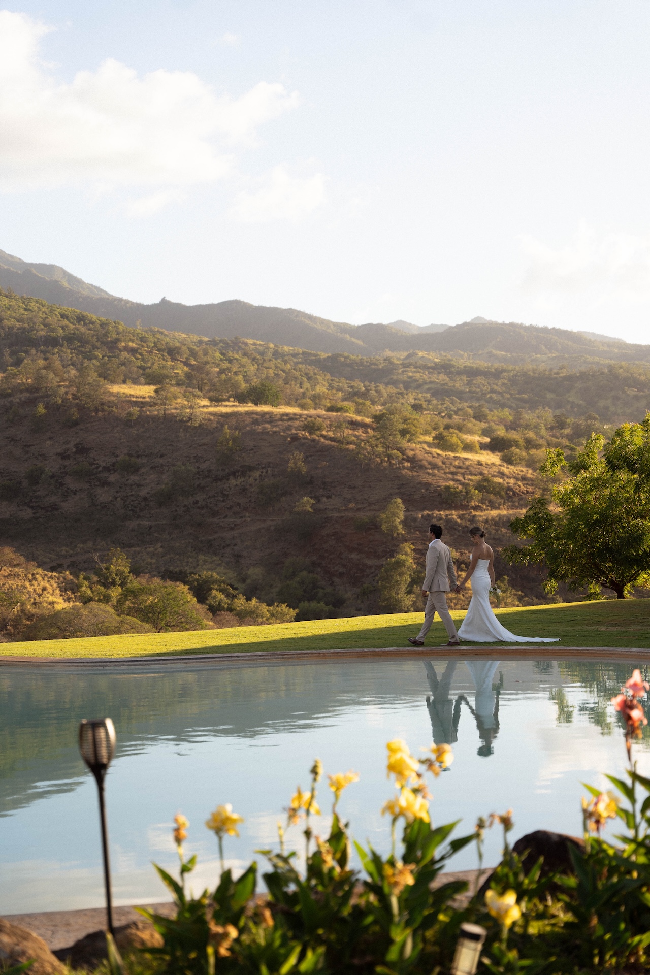 The bride and groom walk side by side near a reflective pool with mountains in the background.