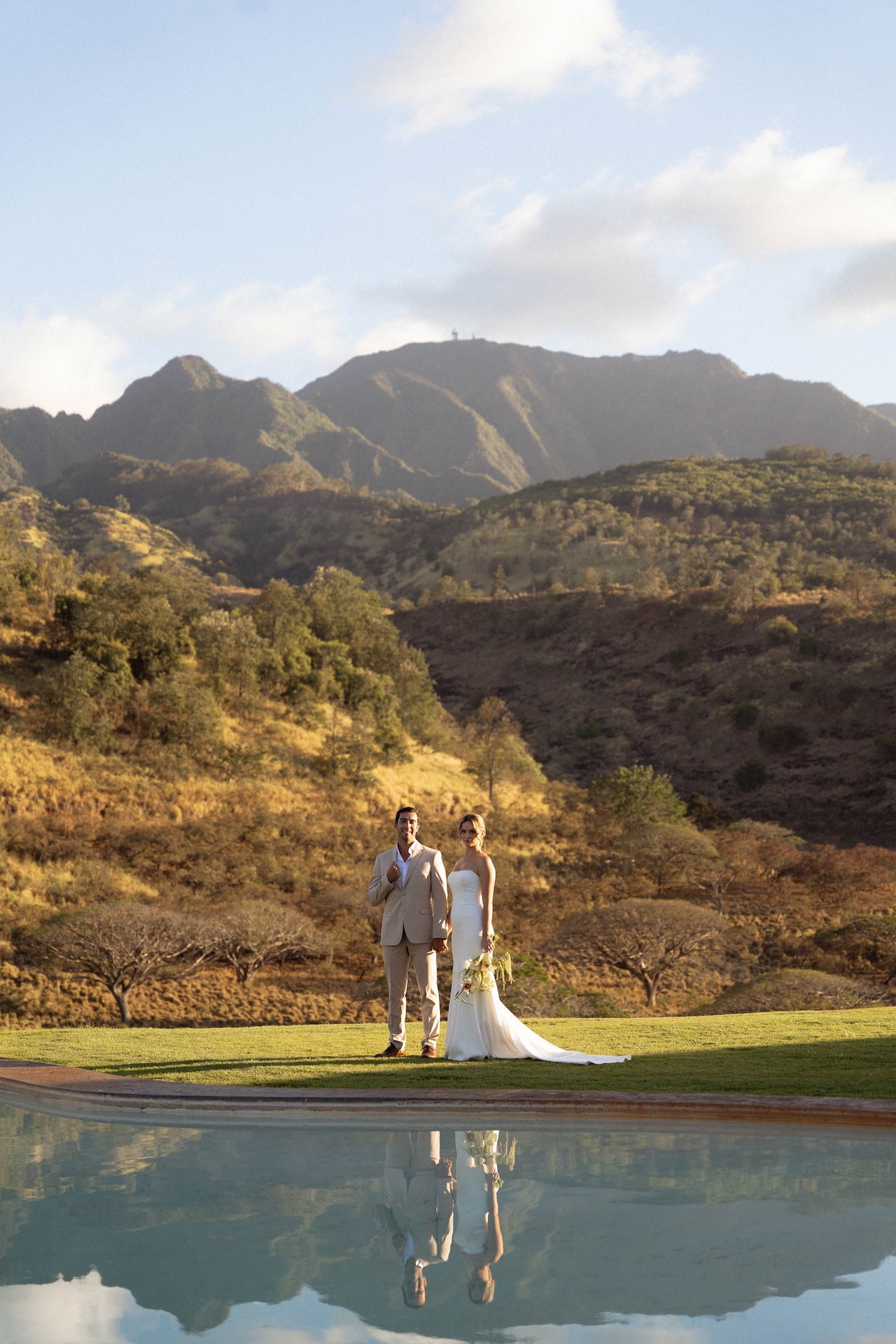 The couple stands hand-in-hand near a pool, their reflections visible in the water, at their Hawaii destination wedding.