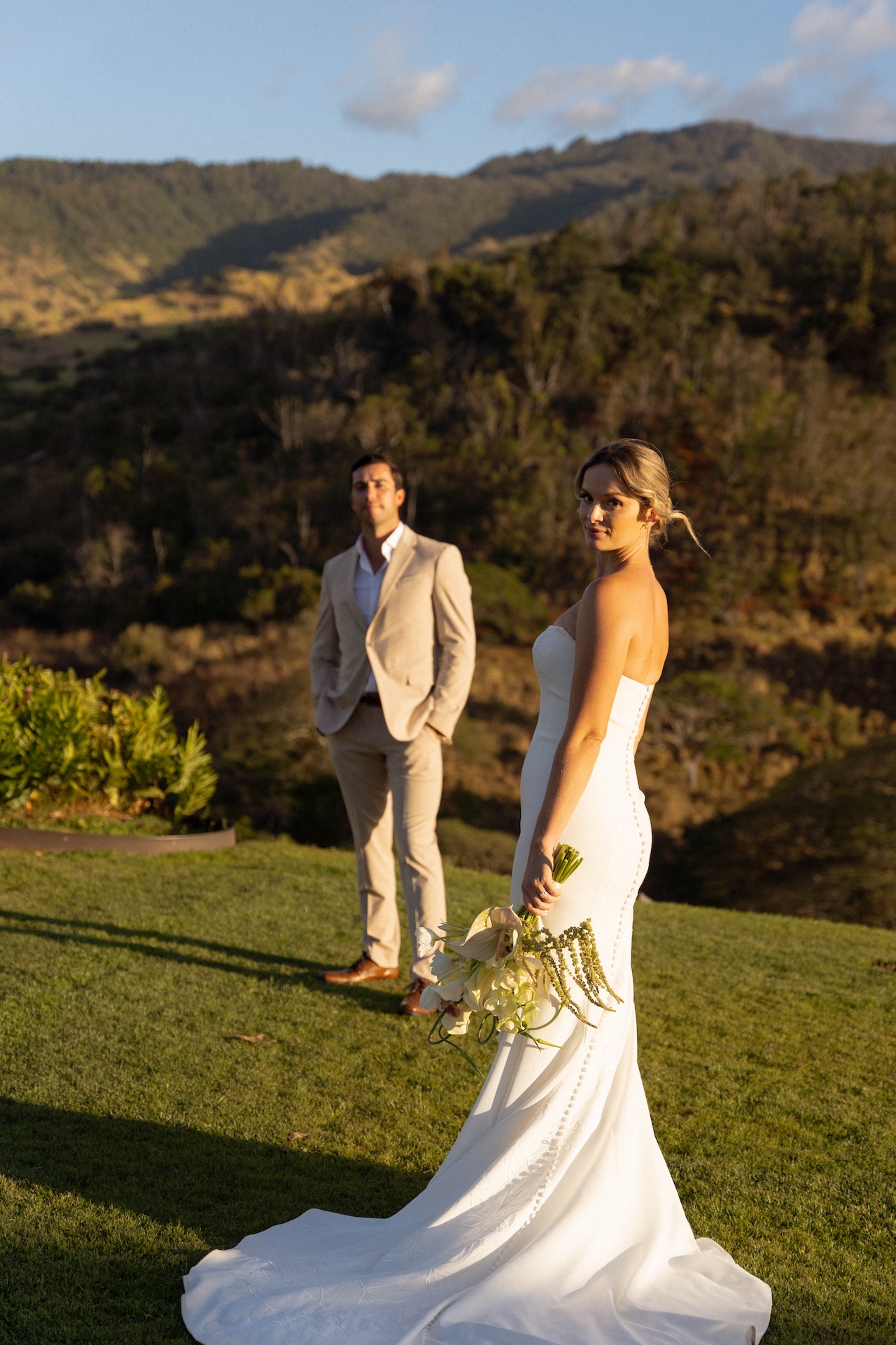 The bride turns toward the camera while the groom stands behind her on a grassy hill at golden hour.