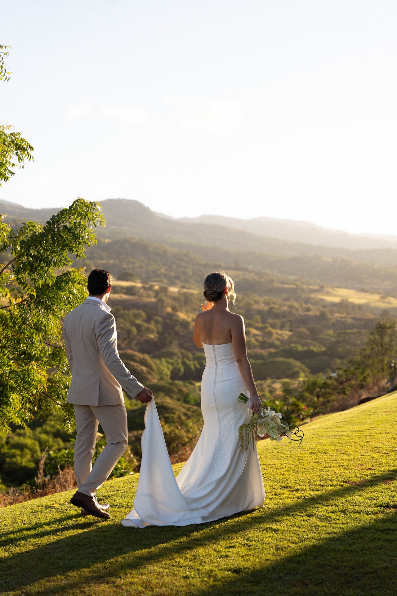 The bride and groom walk together across a grassy hill at sunset with a scenic mountain view at their hawaii destination wedding.
