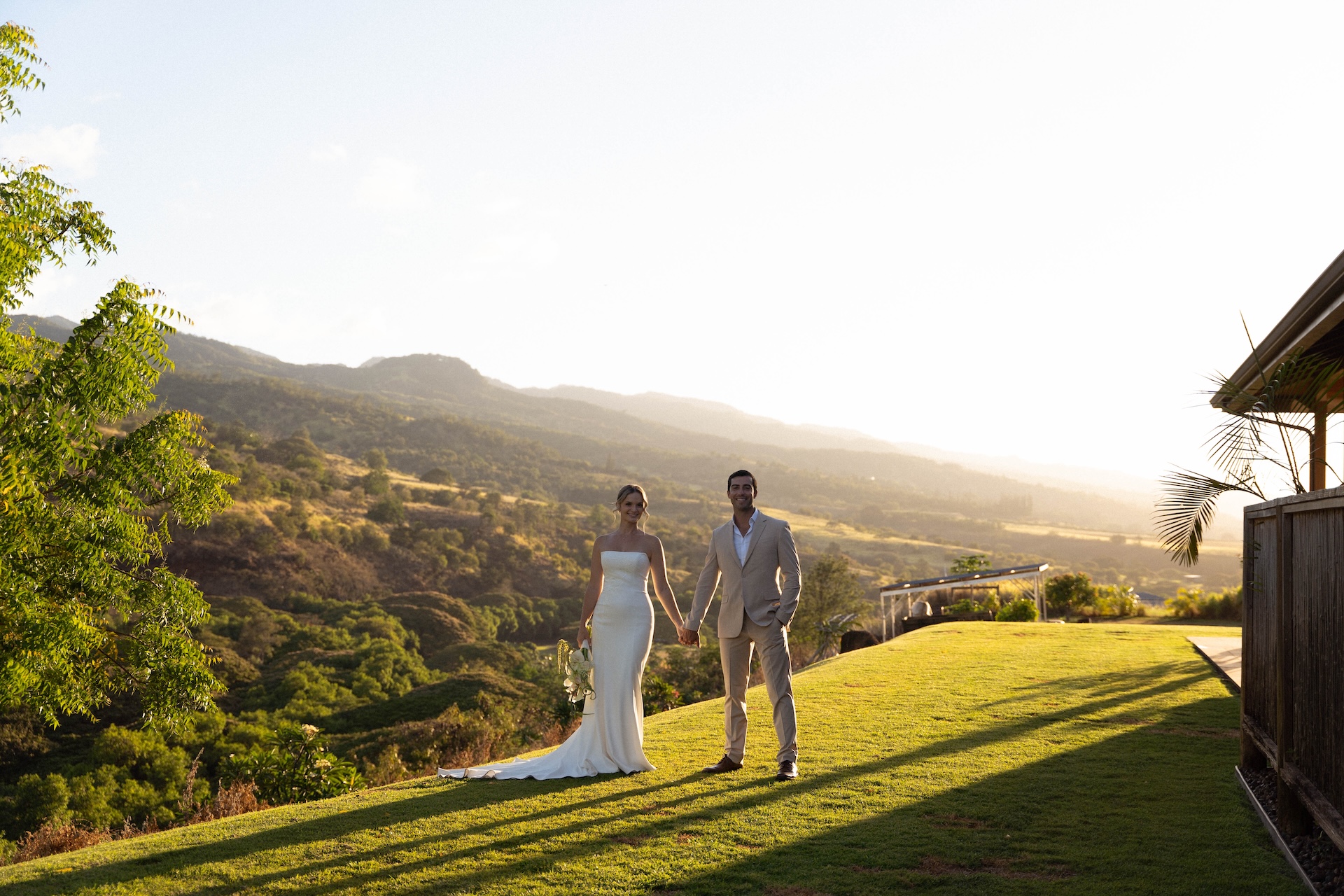 Bride and groom holding hands on a grassy hill at Kaʻala Vista with the mountains glowing in the sunset light.