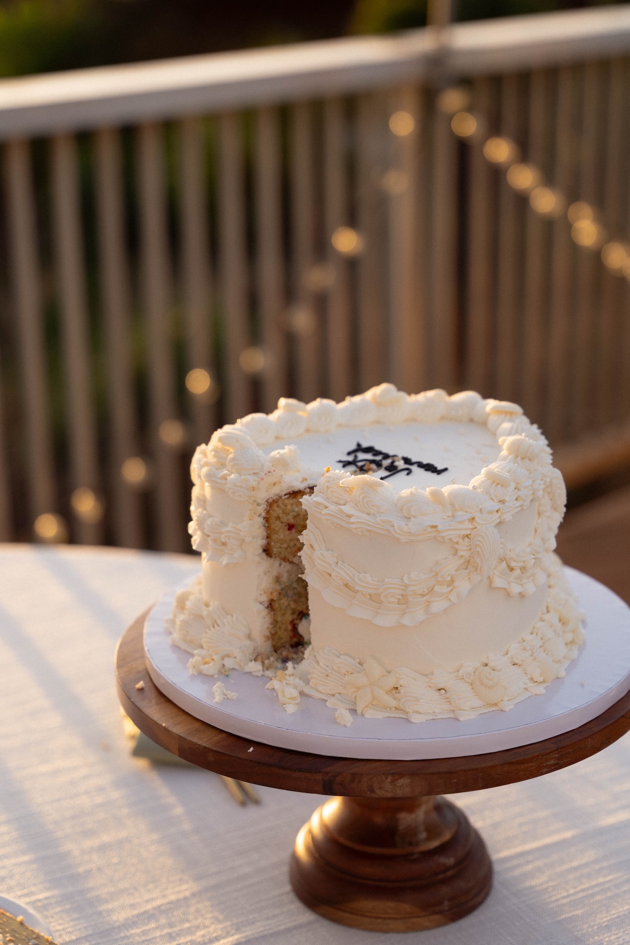 A partially eaten white cake with piped floral icing details sits on a wooden cake stand in the evening light.