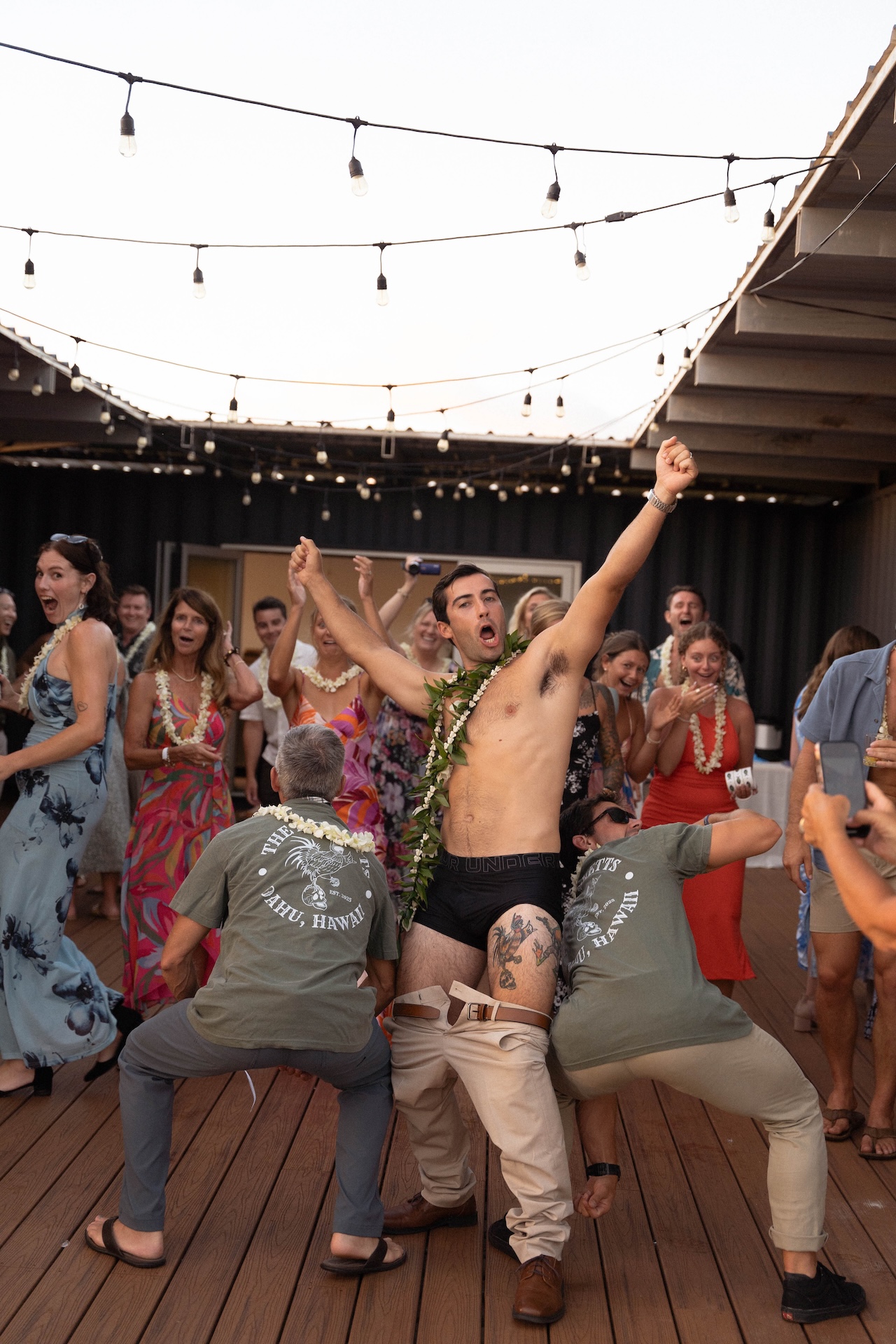 A man in underwear with a lei celebrates on the dance floor surrounded by cheering guests.