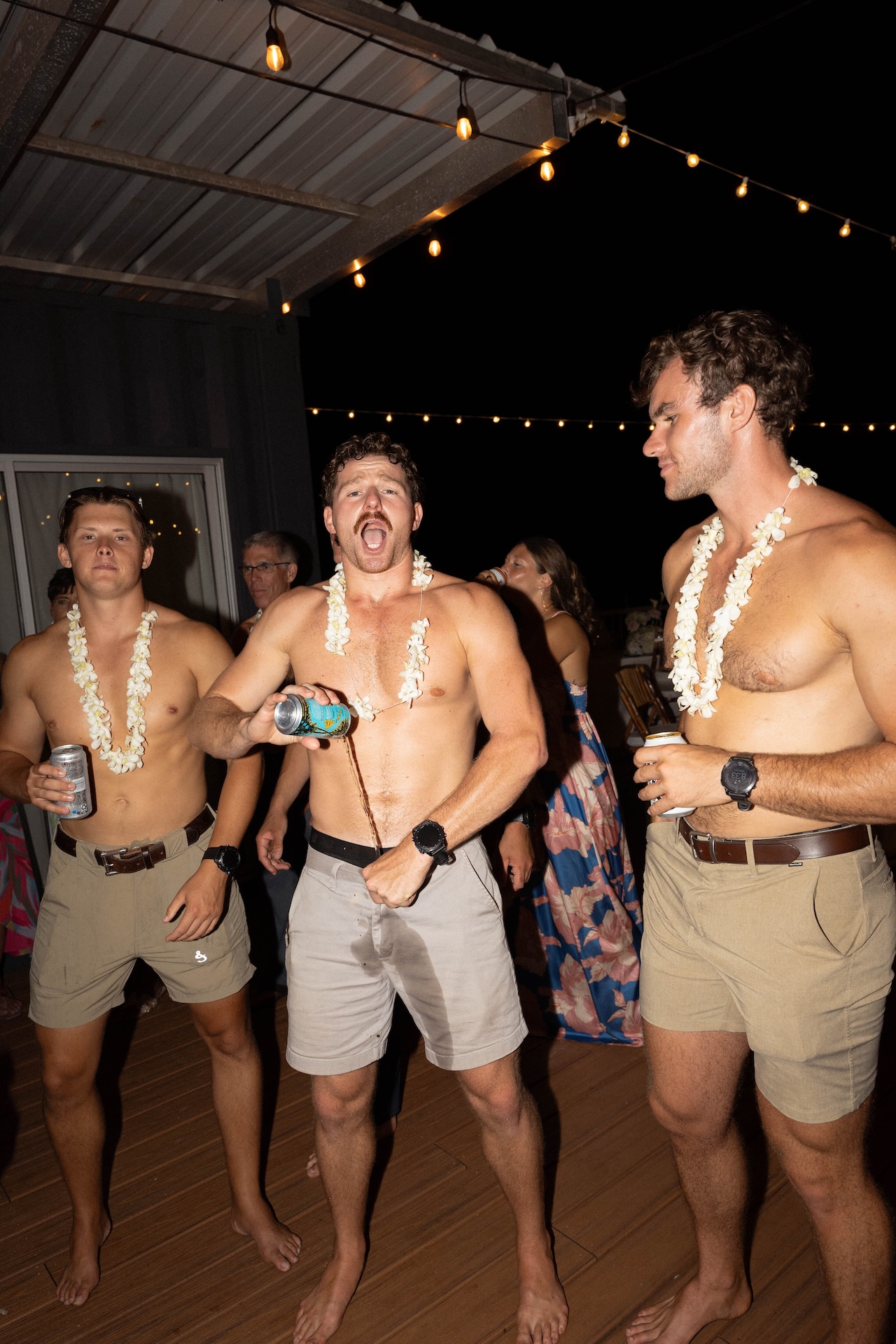 Three shirtless male guests wearing shorts and floral leis dance and drink beer at the wedding reception under string lights.
