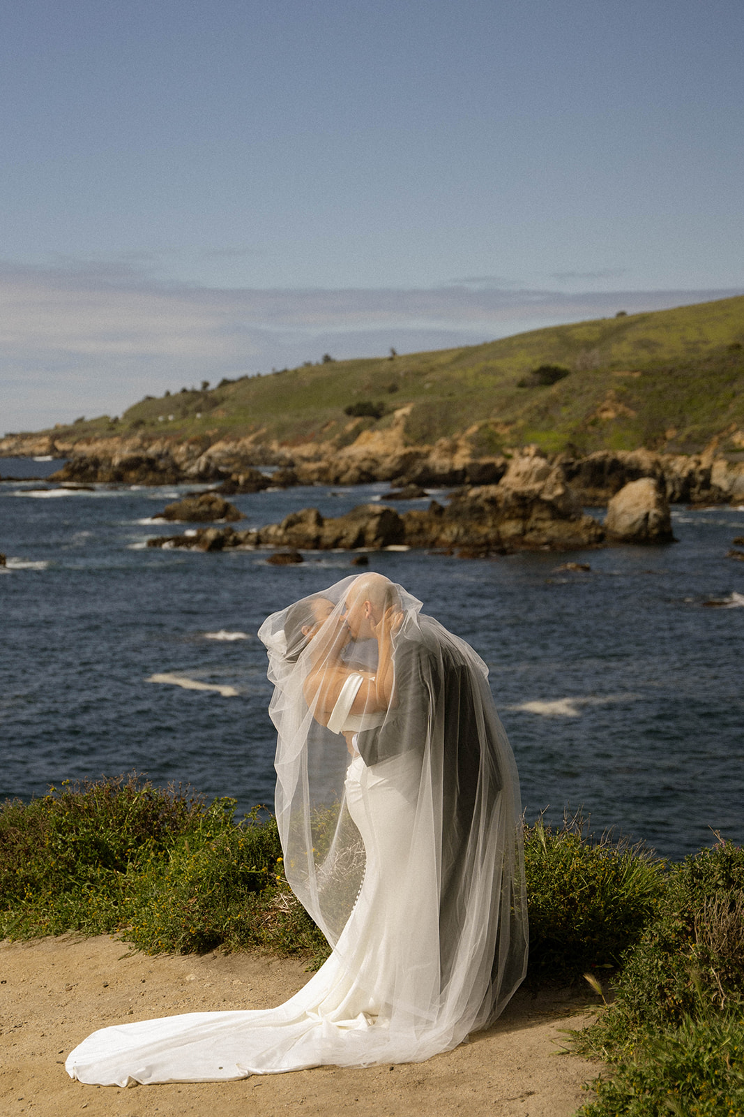 Couple kissing under the bride’s veil with dramatic coastal cliffs behind them during their Big Sur wedding portraits.