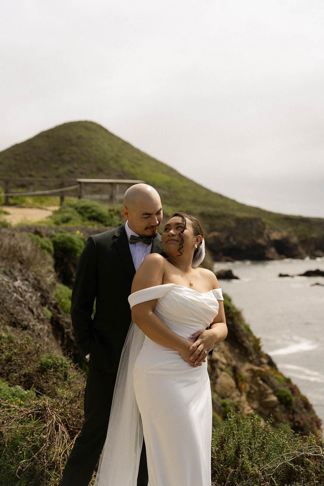 Groom holding bride from behind as they pose on a scenic cliffside during their Big Sur wedding near the coast.