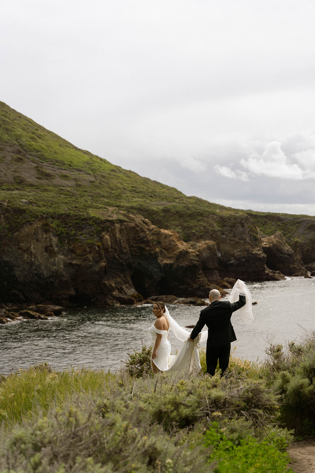 Bride and groom walking along the rugged coastline, with the groom holding the bride’s veil at their Big Sur wedding.