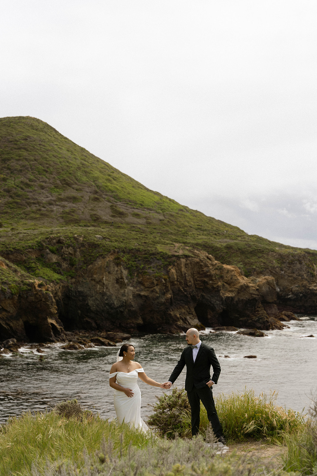 Bride and groom holding hands on a cliffside overlooking the ocean at their Big Sur wedding.