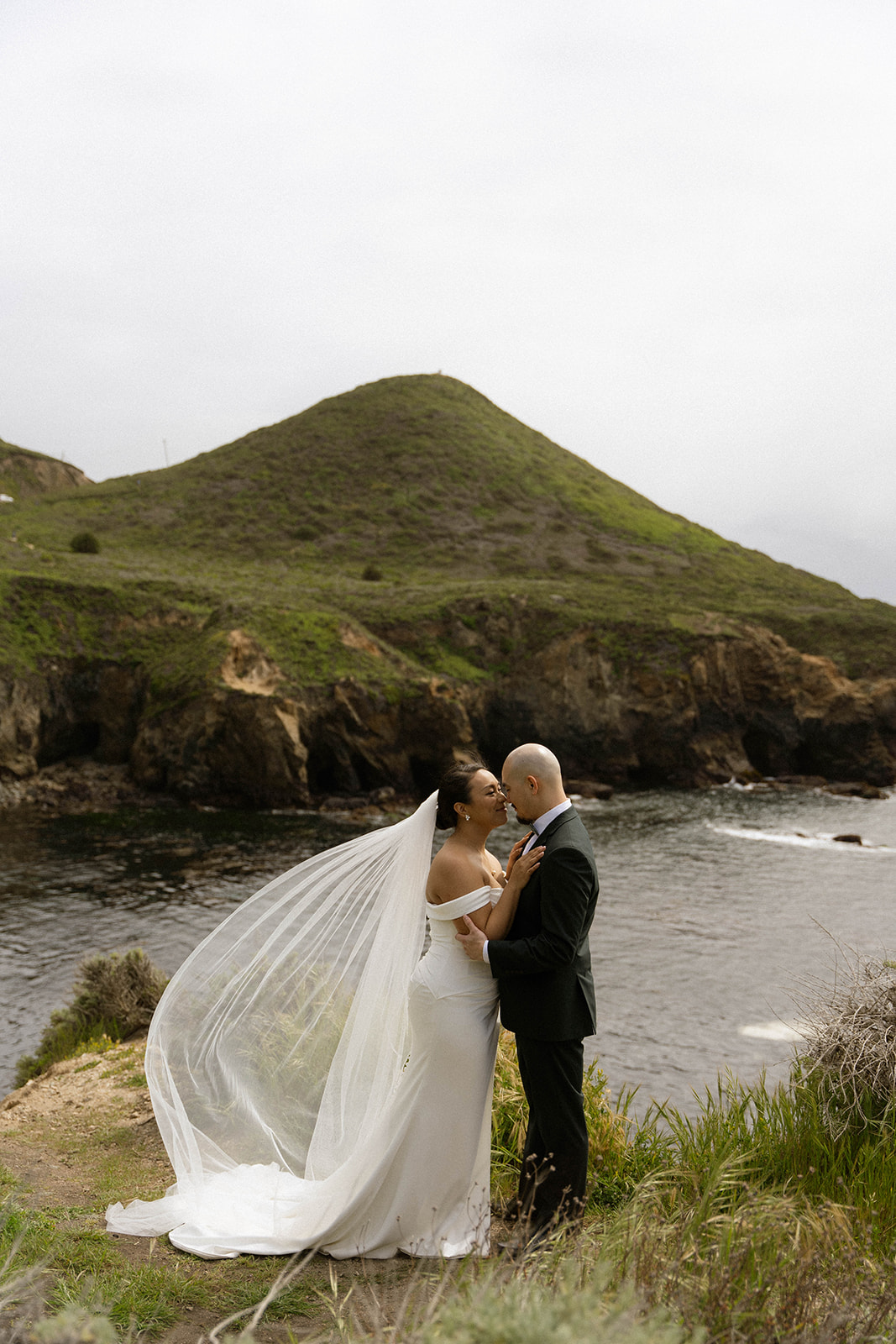 Bride’s veil blowing in the wind as the couple embraces on the cliffs at their Big Sur wedding venue overlooking the ocean.