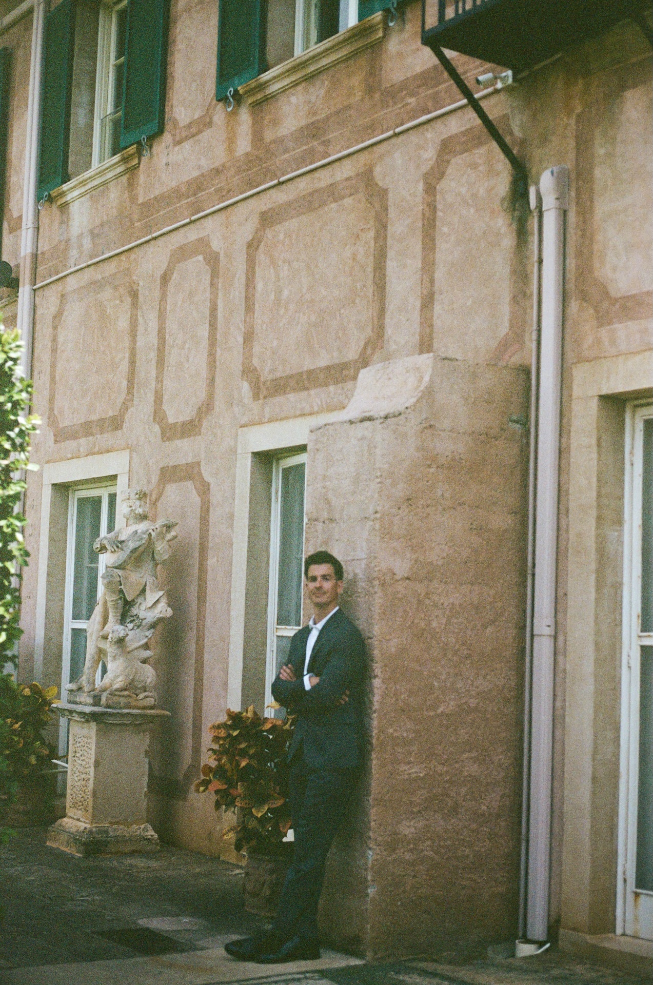 A full-length portrait of the groom leaning casually against a textured wall with green shutters and a sculpture nearby.