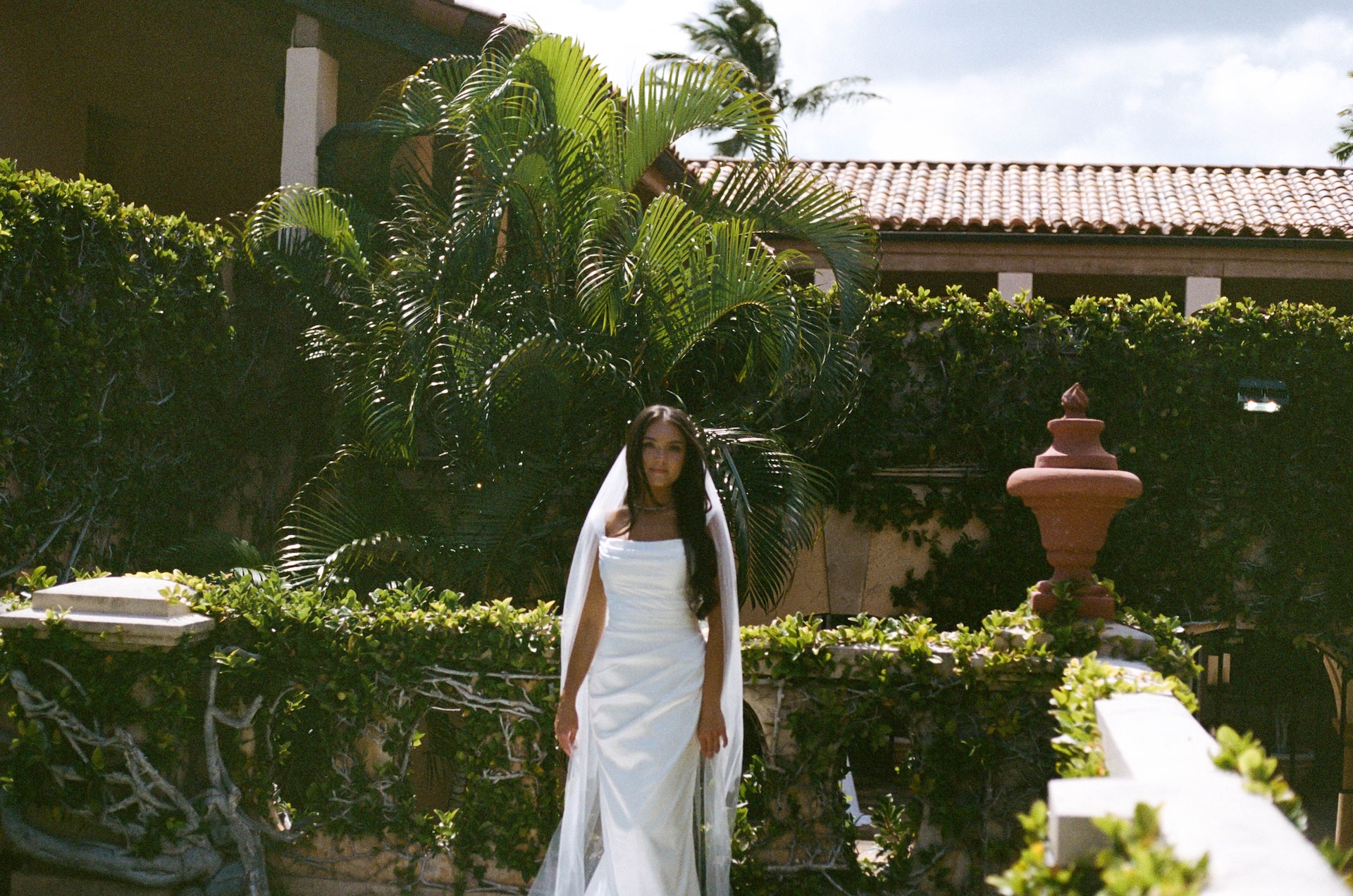 The bride stands in front of a Mediterranean-style villa surrounded by lush palms, her veil flowing in the breeze.