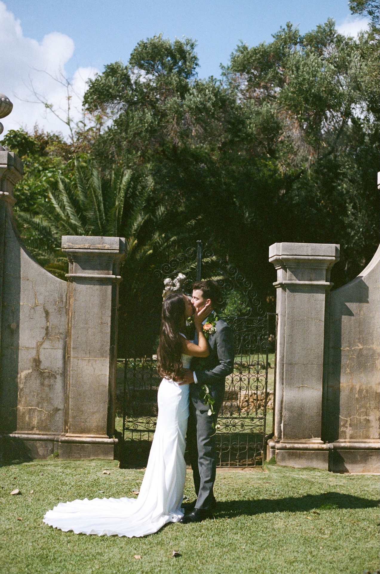 The couple kisses in front of a wrought iron gate framed by stone columns, her dress fanned out perfectly on the grass.