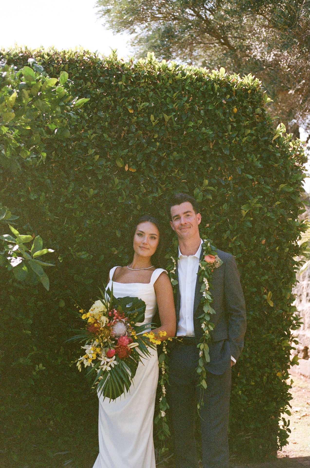 The bride and groom stand smiling against a manicured hedge, the bride holding a vibrant bouquet of tropical florals — a joyful moment by a destination wedding photographer.