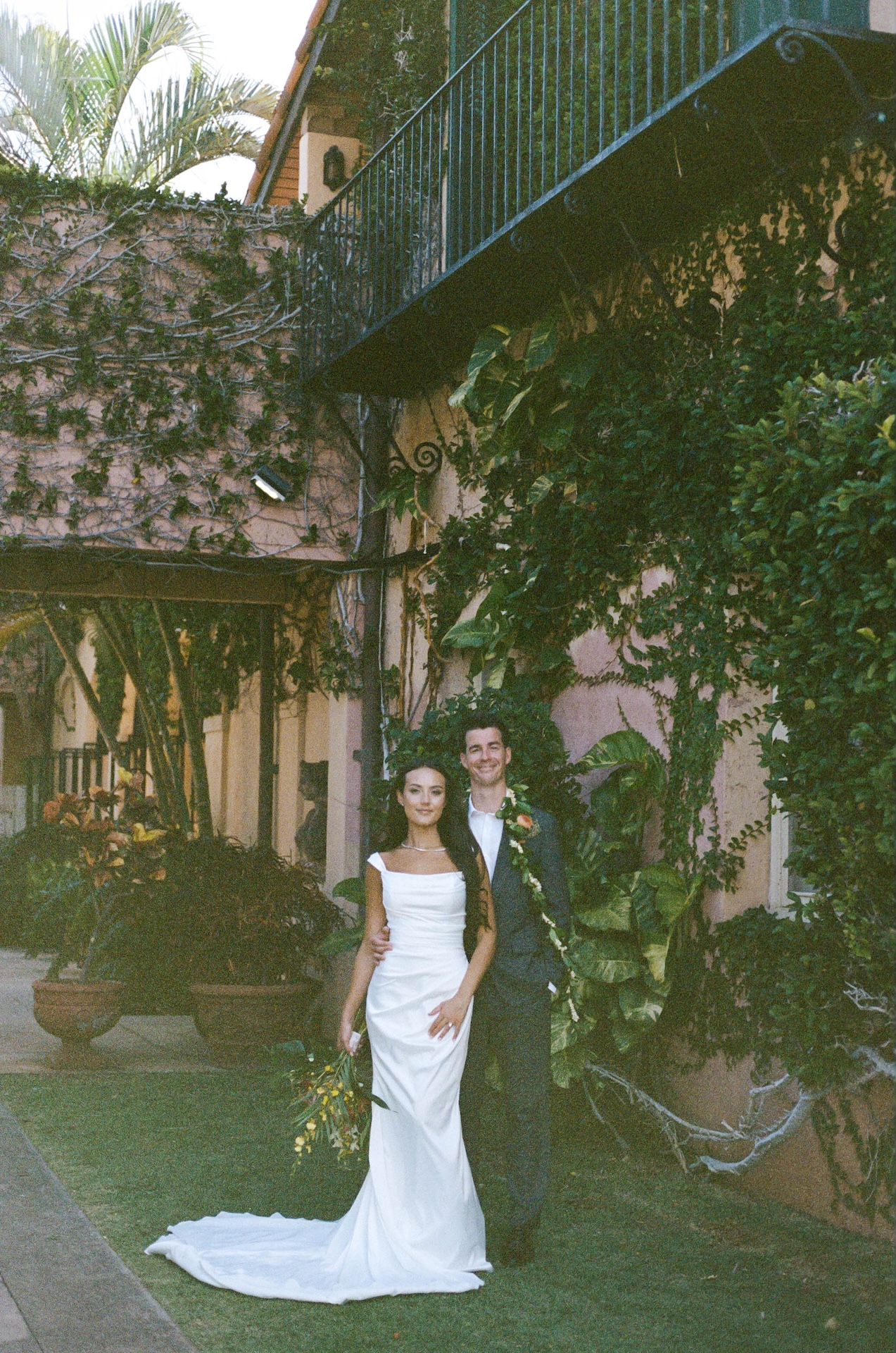 The bride and groom pose in front of a vine-covered villa wall, surrounded by tropical greenery in soft, natural light.