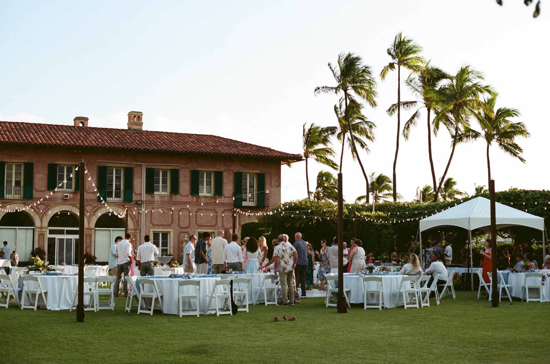 Guests mingle under string lights on a lawn set for a reception, with round tables, a pink villa, and swaying palm trees in the background.