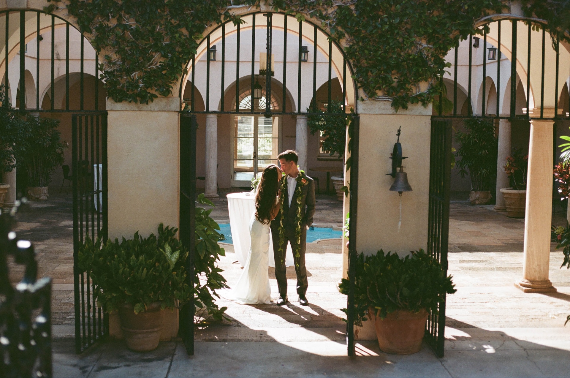 The couple shares a soft kiss beneath a grand arched gate covered in vines, framed by symmetrical pillars and sunlit courtyards.