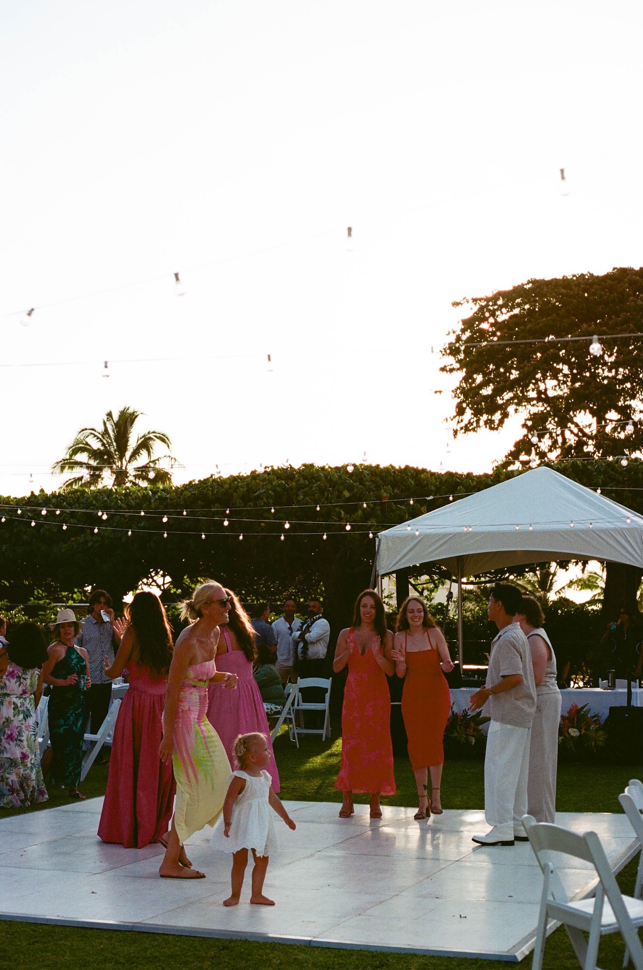 A joyful group of wedding guests and a barefoot toddler dance together on a white dance floor during golden hour.