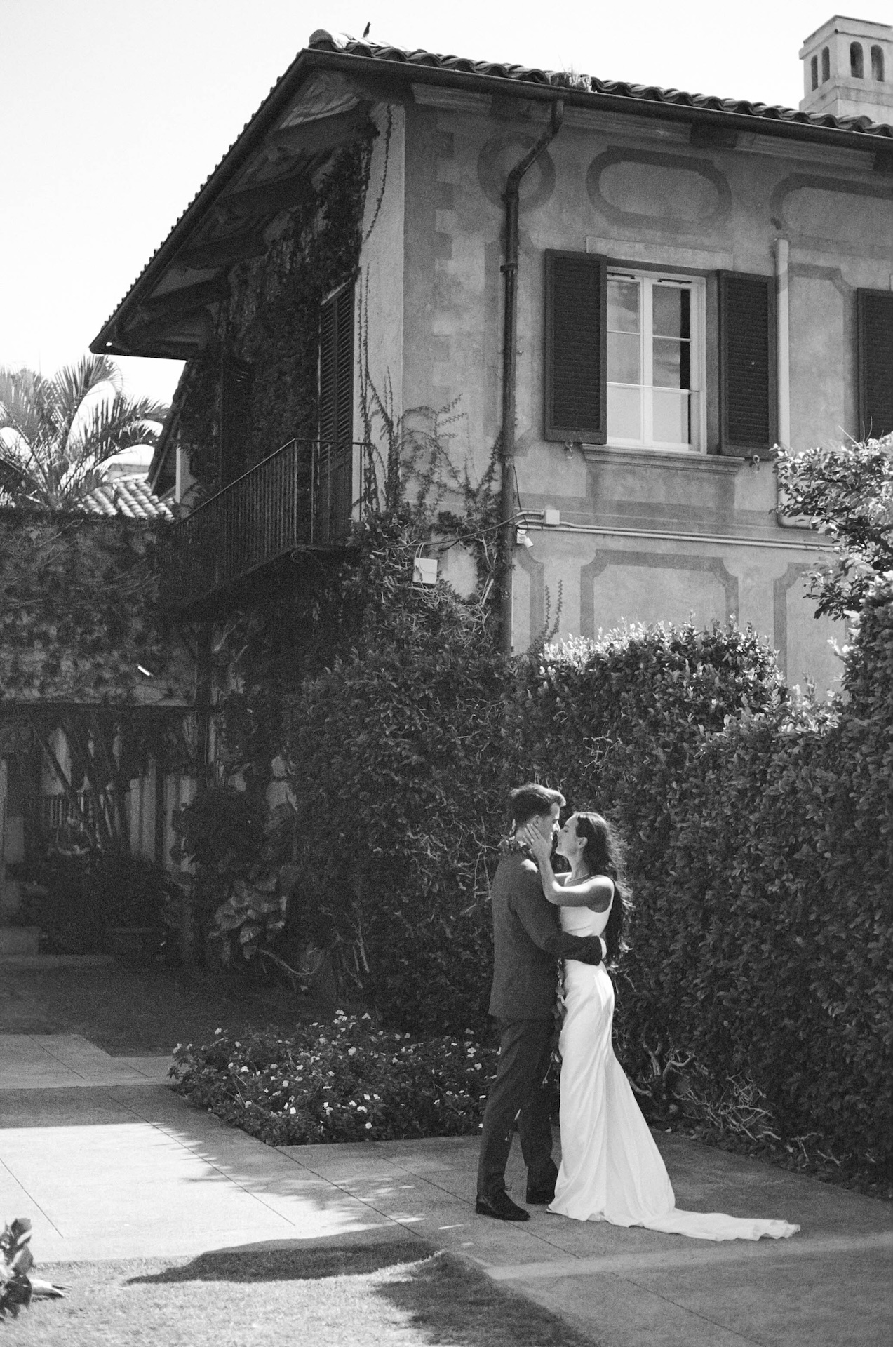 Black and white photo of a bride and groom embracing on a sunlit path in front of a Mediterranean-style villa covered in ivy, surrounded by lush hedges — a timeless moment captured by a destination wedding photographer.